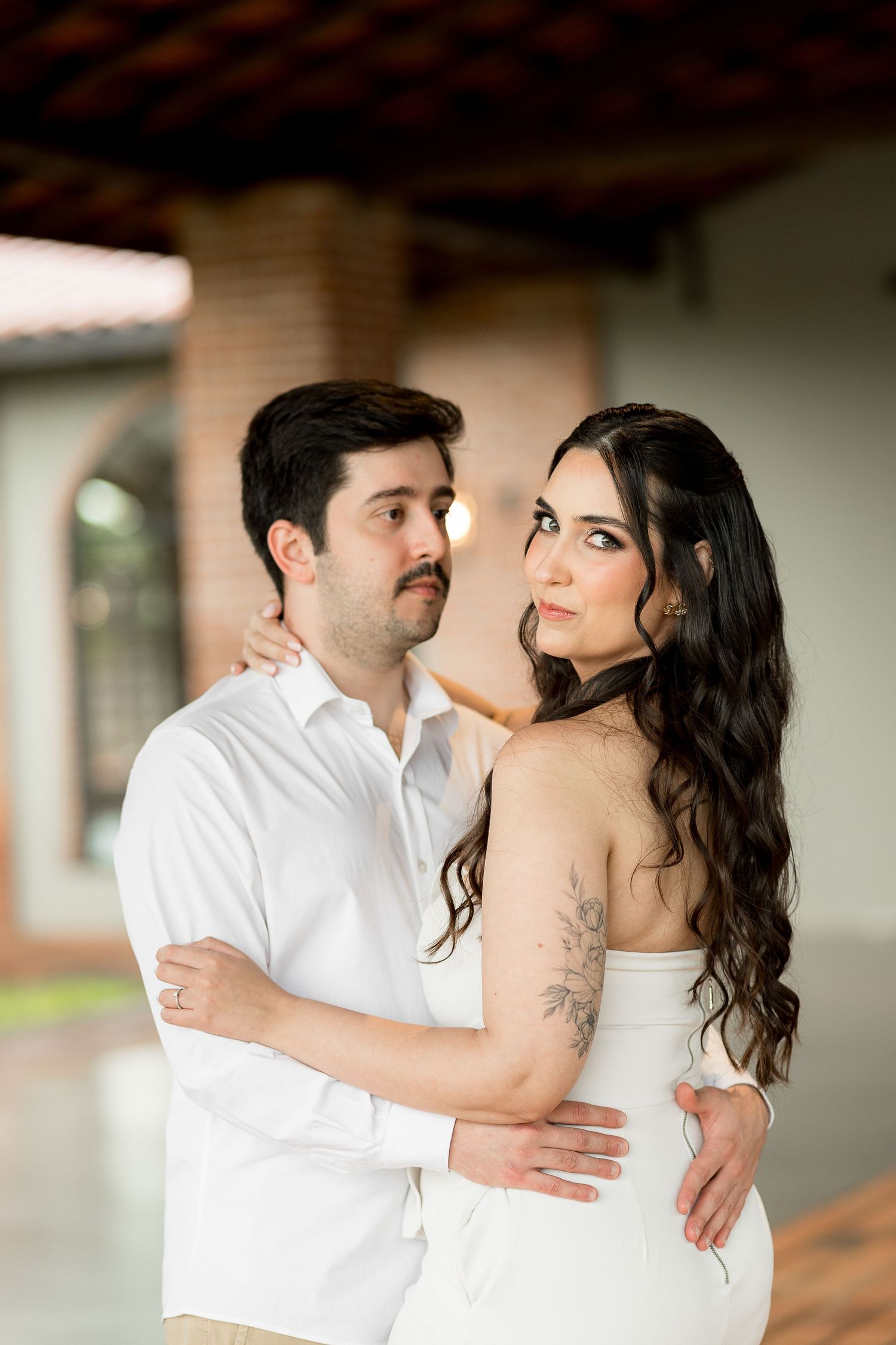 Couple in white outfits embracing outdoors, woman with long dark wavy hair and floral arm tattoo gazing away romantically.