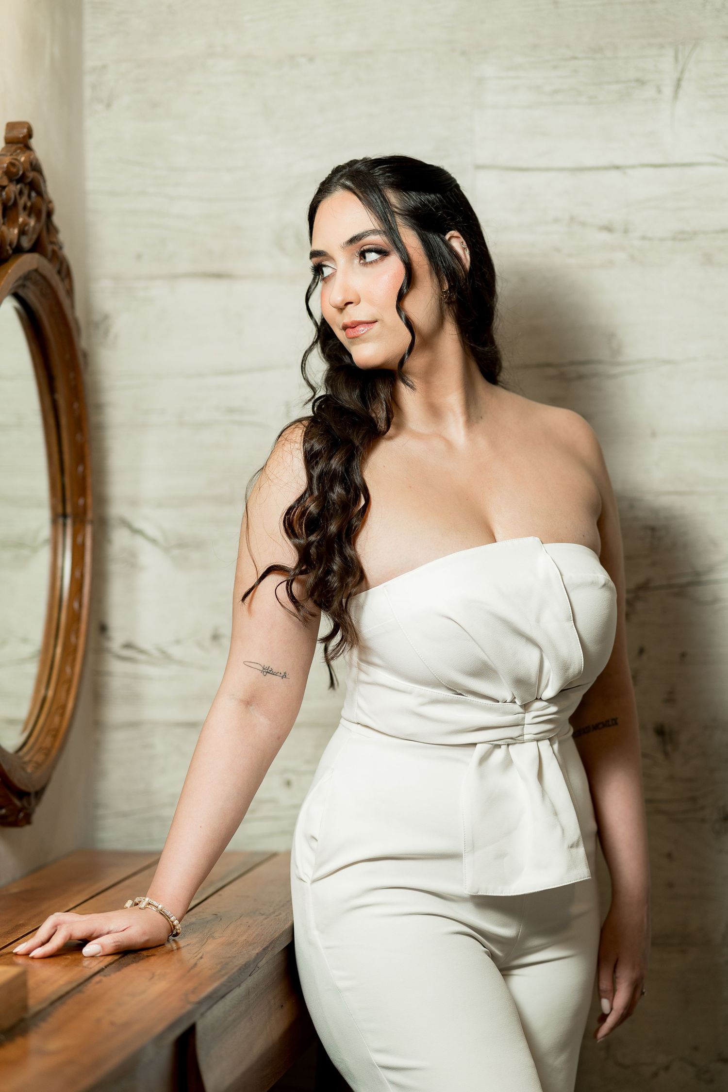 Woman in white strapless jumpsuit with curly dark hair posing elegantly beside a vintage mirror.