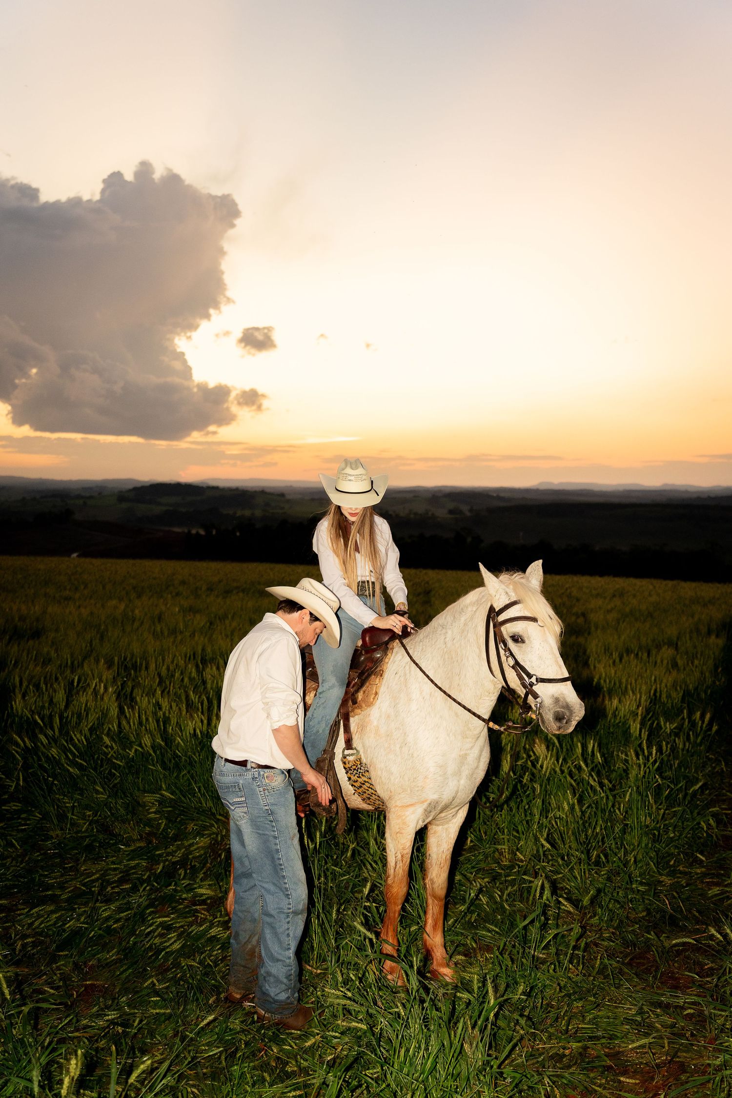 Couple in cowboy hats with a white horse in a green field at sunset, romantic western countryside scene.