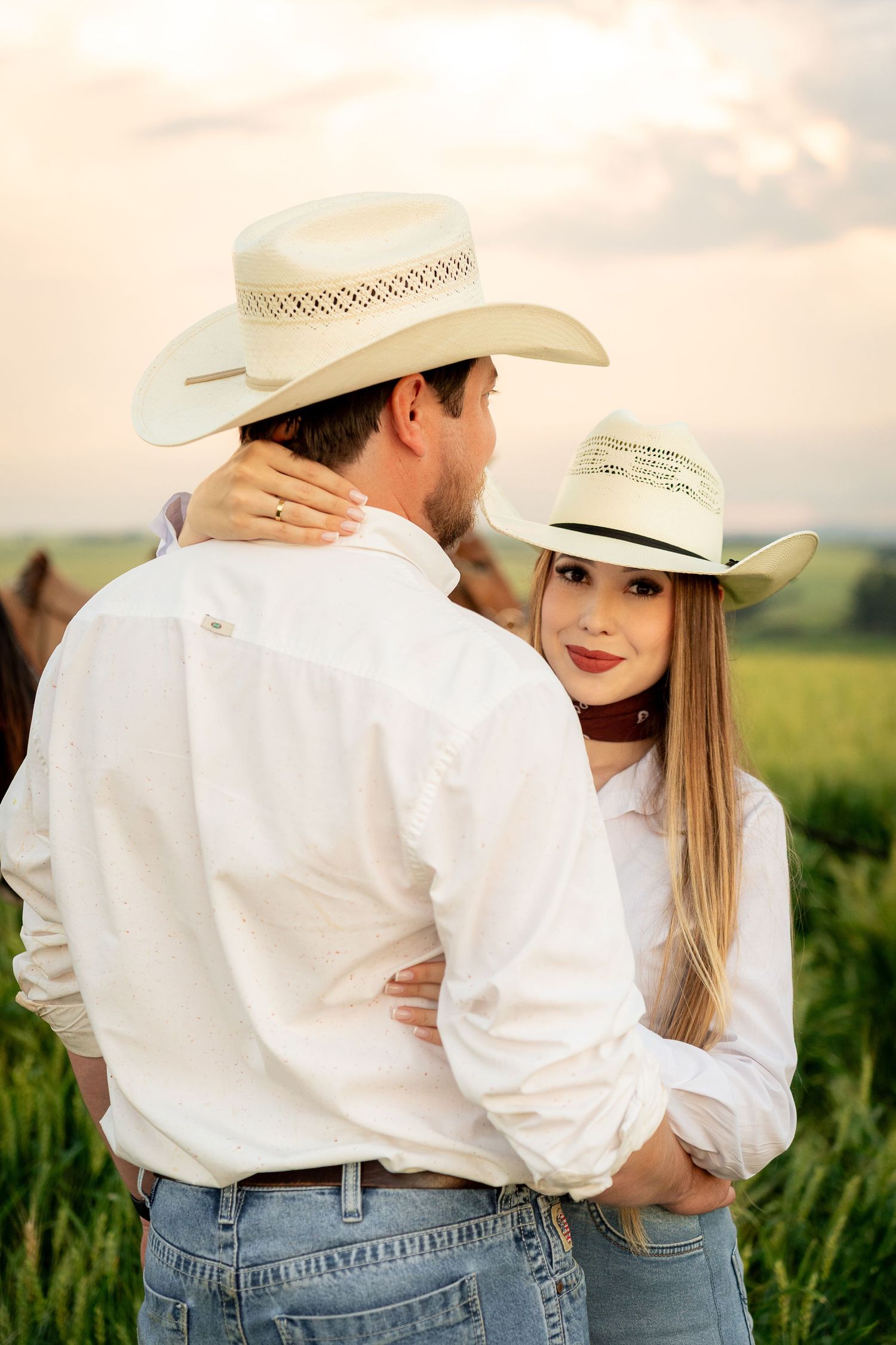 A couple in white shirts and cowboy hats embrace in a green field at sunset, with a horse visible behind them.