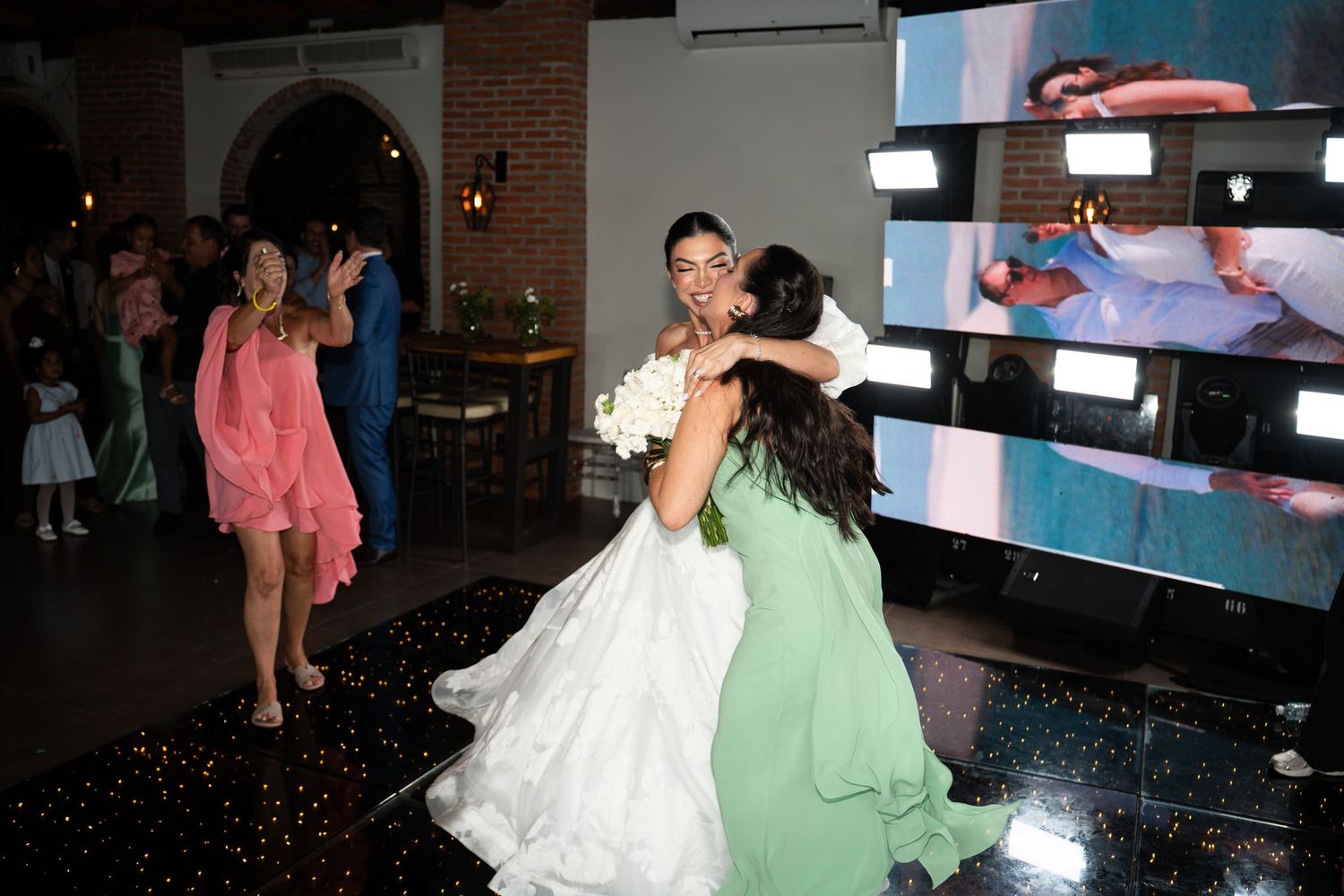 Bride in white gown hugging guest in green dress on sparkly dance floor at elegant wedding reception.