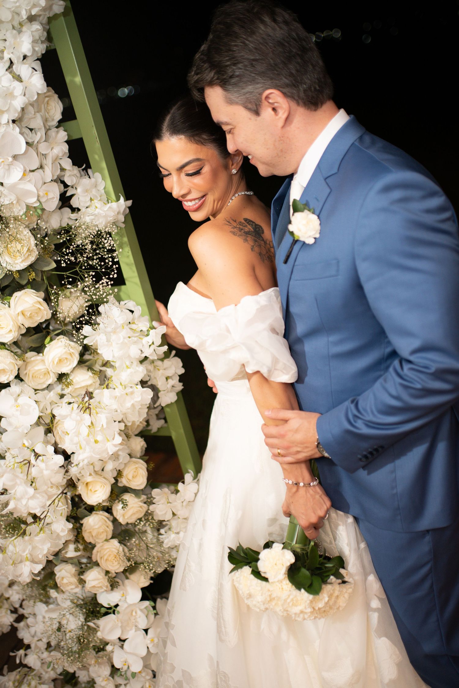 Bride in white off-shoulder gown and groom in blue suit embrace near white floral arch at elegant wedding ceremony.