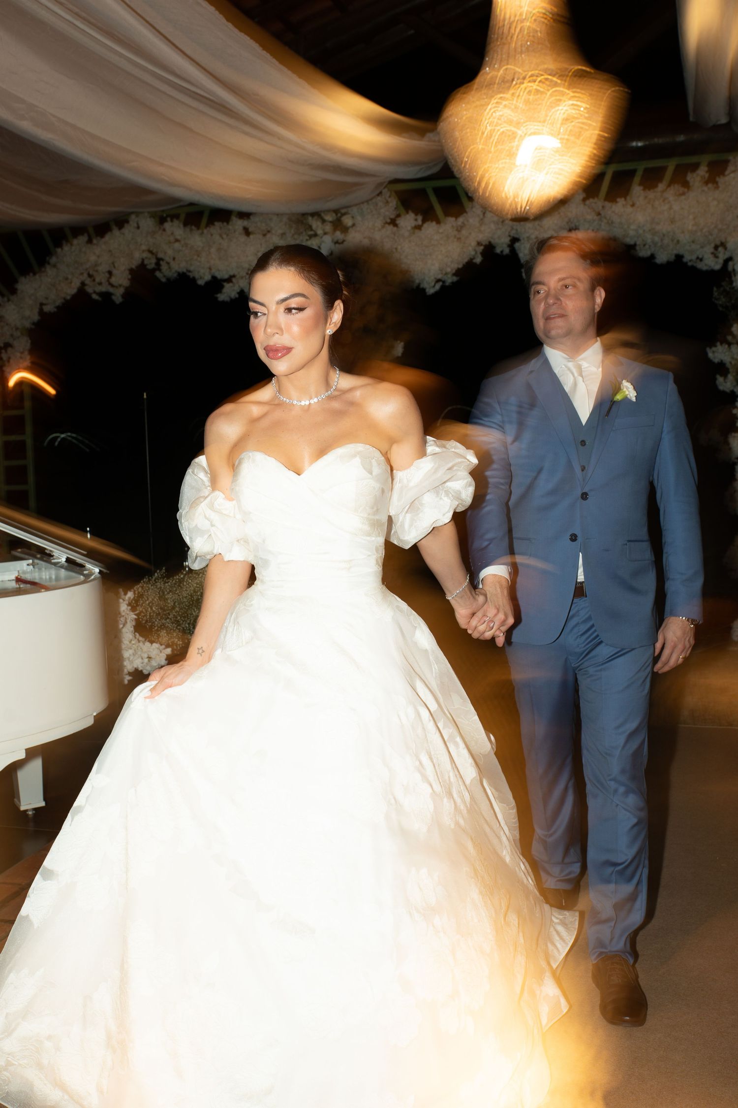 Bride in white ball gown and groom in blue suit hold hands during their elegant outdoor wedding reception at night.
