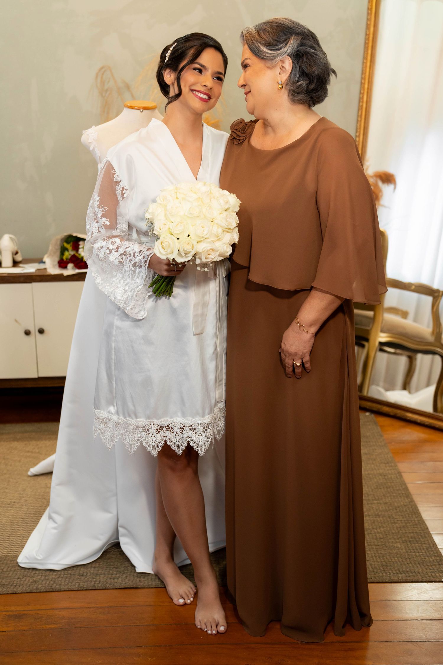 Bride in white lace robe holding bouquet of white roses stands with older woman in brown dress indoors.