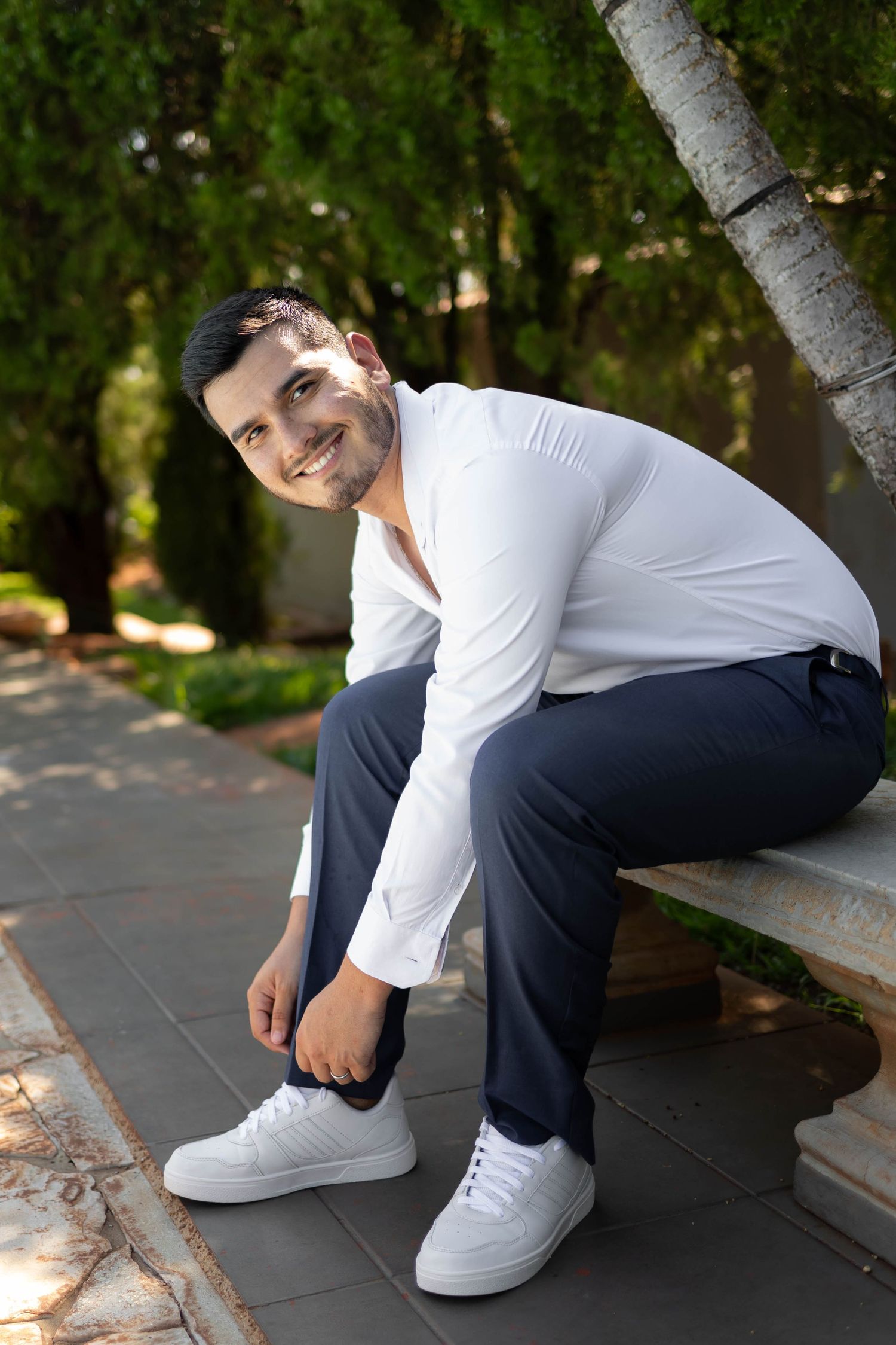 Smiling man in white shirt and navy pants sits on stone bench in lush green park, wearing white sneakers.