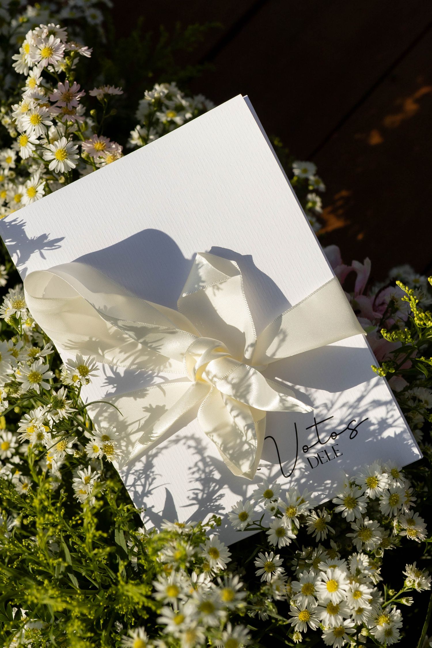 White wedding vow book with cream ribbon bow surrounded by delicate white daisy flowers in natural sunlight.
