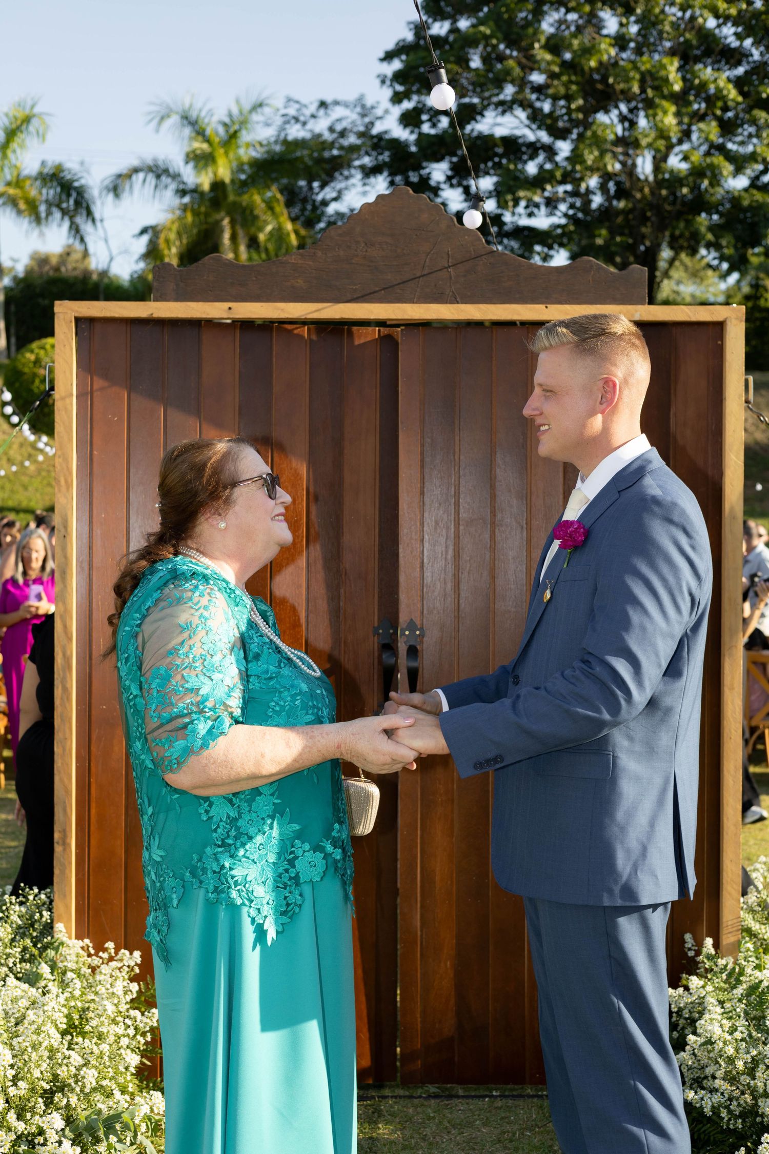 Groom in blue suit holds hands with woman in teal lace dress at outdoor wedding ceremony by wooden backdrop.