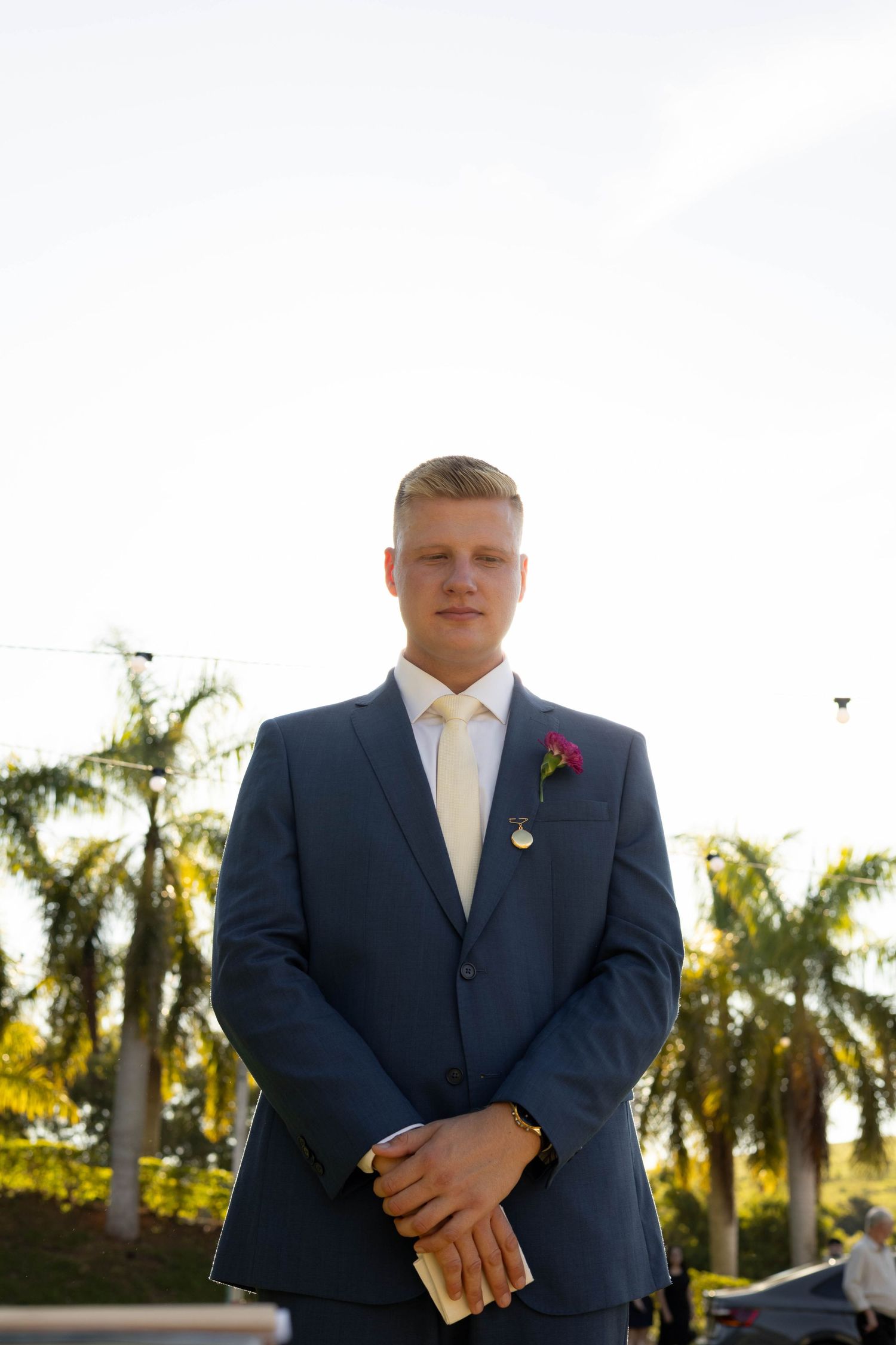 Young groom in navy blue suit with boutonniere holding a book outdoors with palm trees in background.