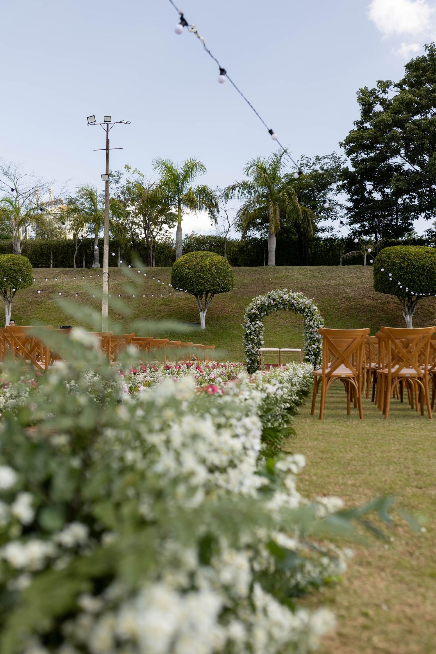 Outdoor wedding ceremony setup with floral arch, wooden chairs, white flowers, string lights, and lush greenery under a clear sky.