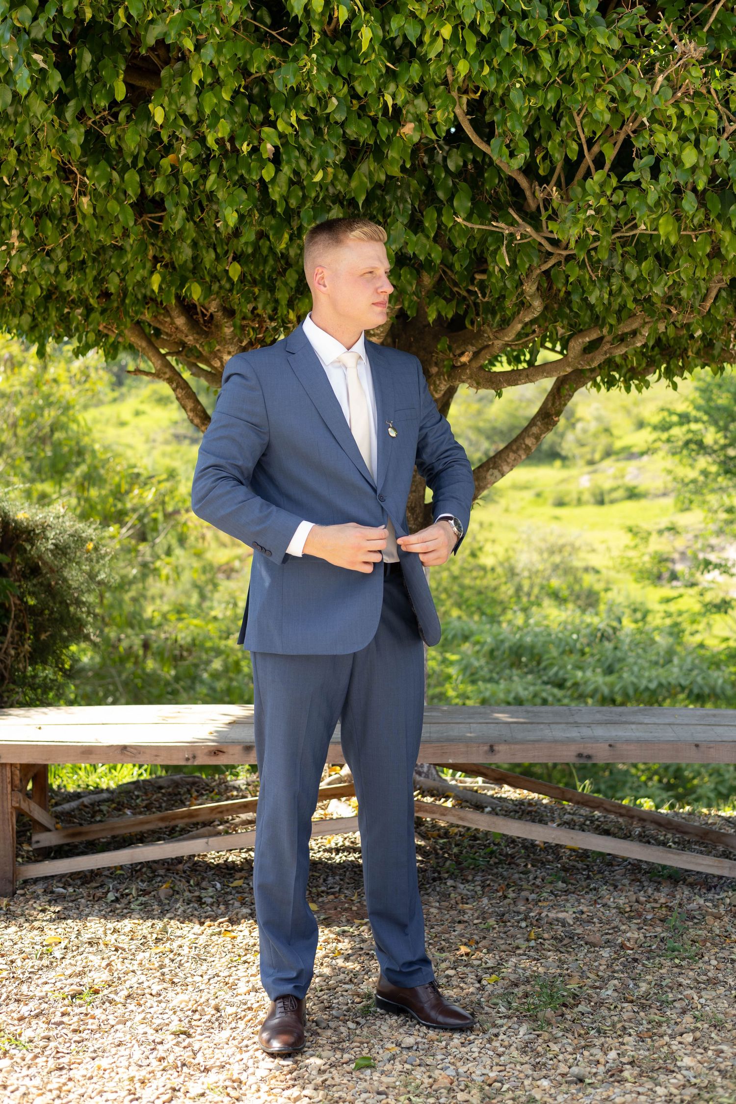 Man in navy blue suit buttoning jacket outdoors near tree, ideal for wedding or formal event attire.