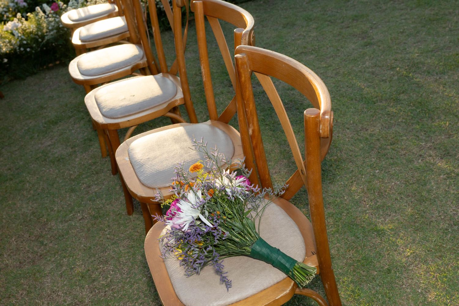 Wooden cross-back chairs arranged in a row on green grass with a colorful wildflower bouquet resting on one seat.