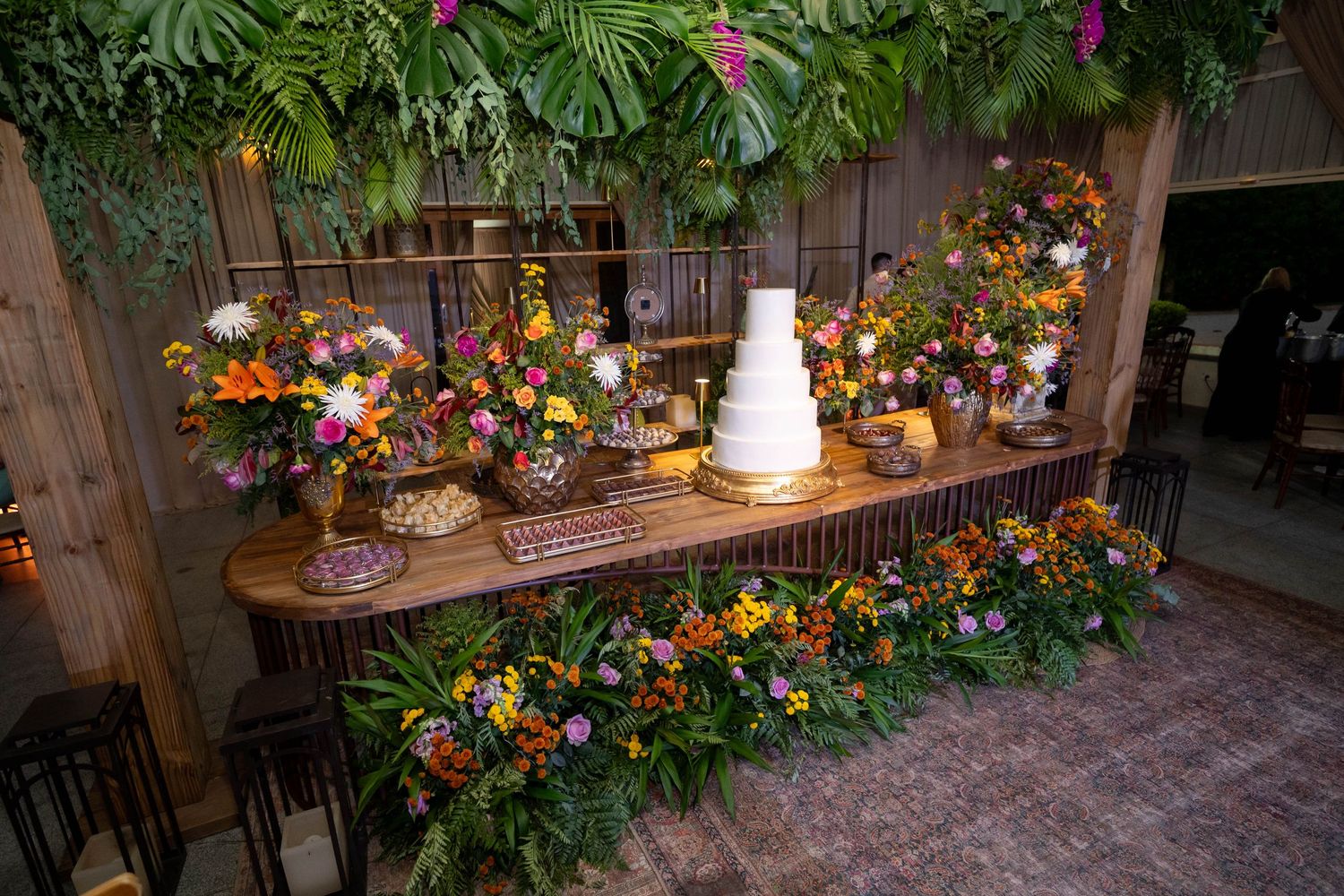 Lush floral wedding dessert table with white tiered cake, colorful flower arrangements, and tropical greenery backdrop.