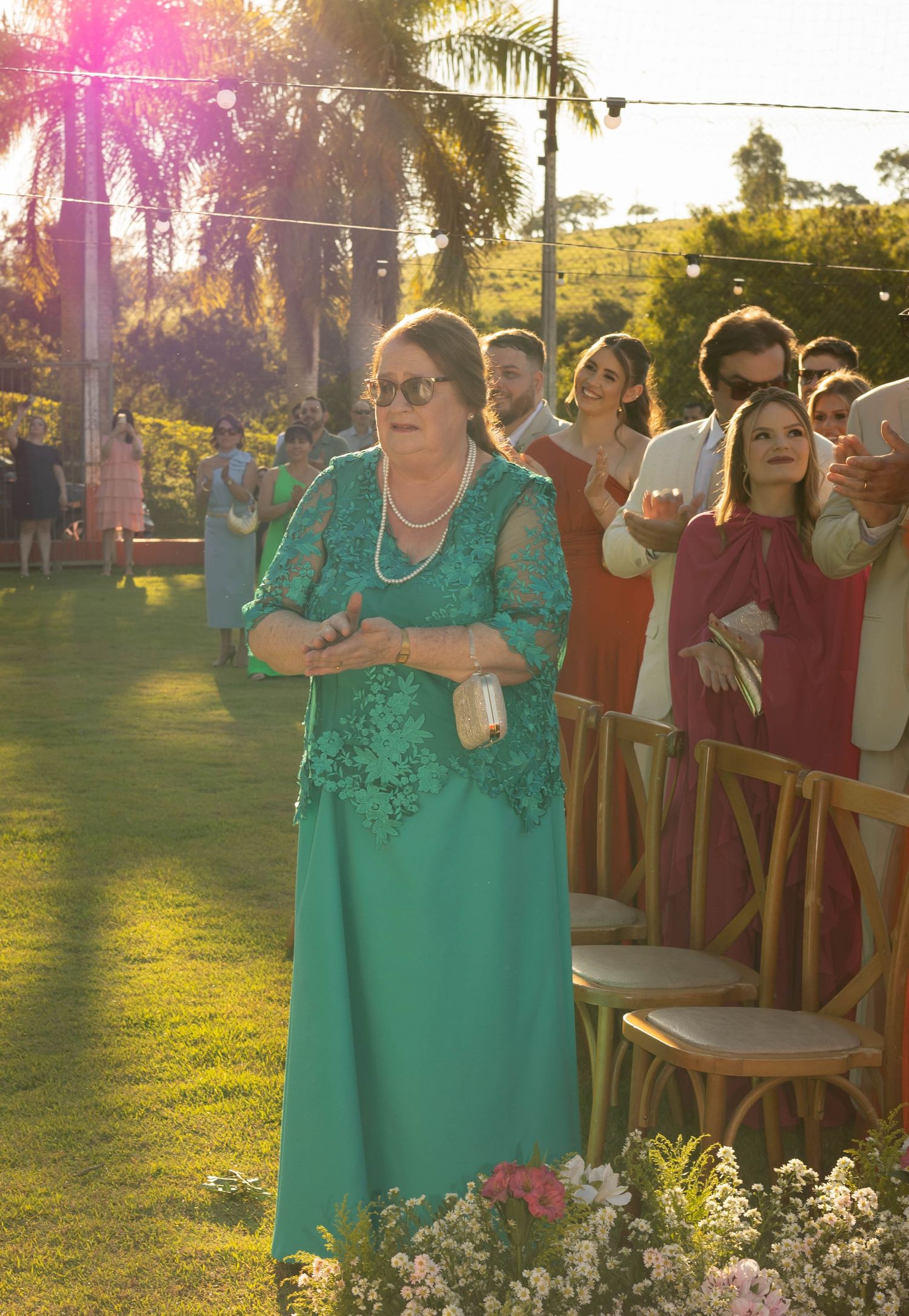 Elegant woman in teal lace gown with pearl necklace at outdoor wedding ceremony on green lawn.