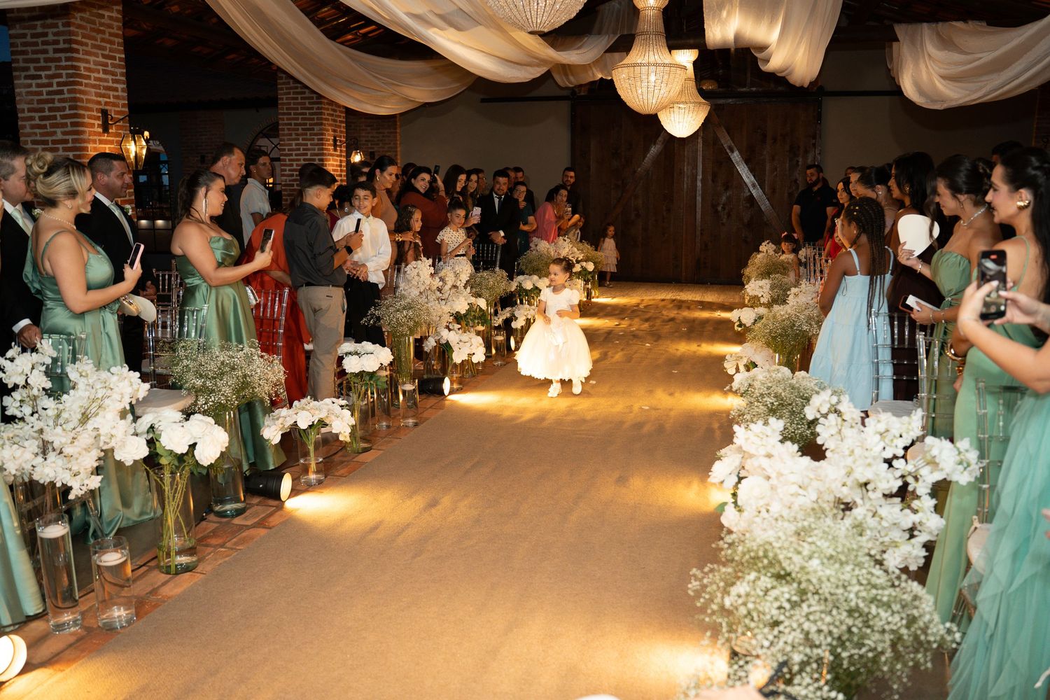 A flower girl walks down a floral-lined wedding aisle surrounded by guests and bridesmaids in green dresses.