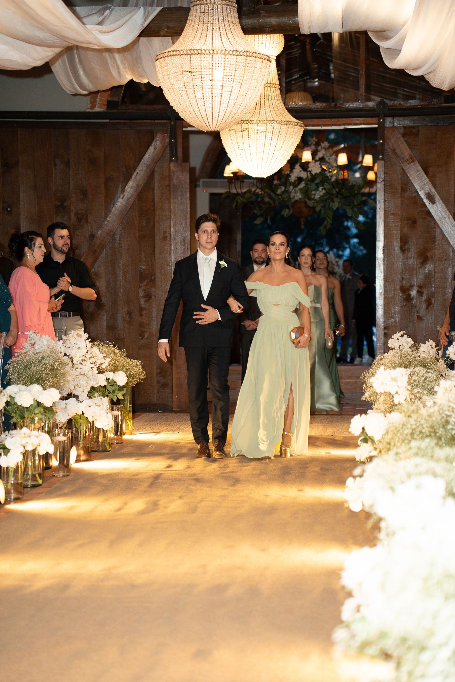 Elegant wedding processional in a barn venue with chandelier lighting, floral arrangements, and guests watching a couple walk the aisle.