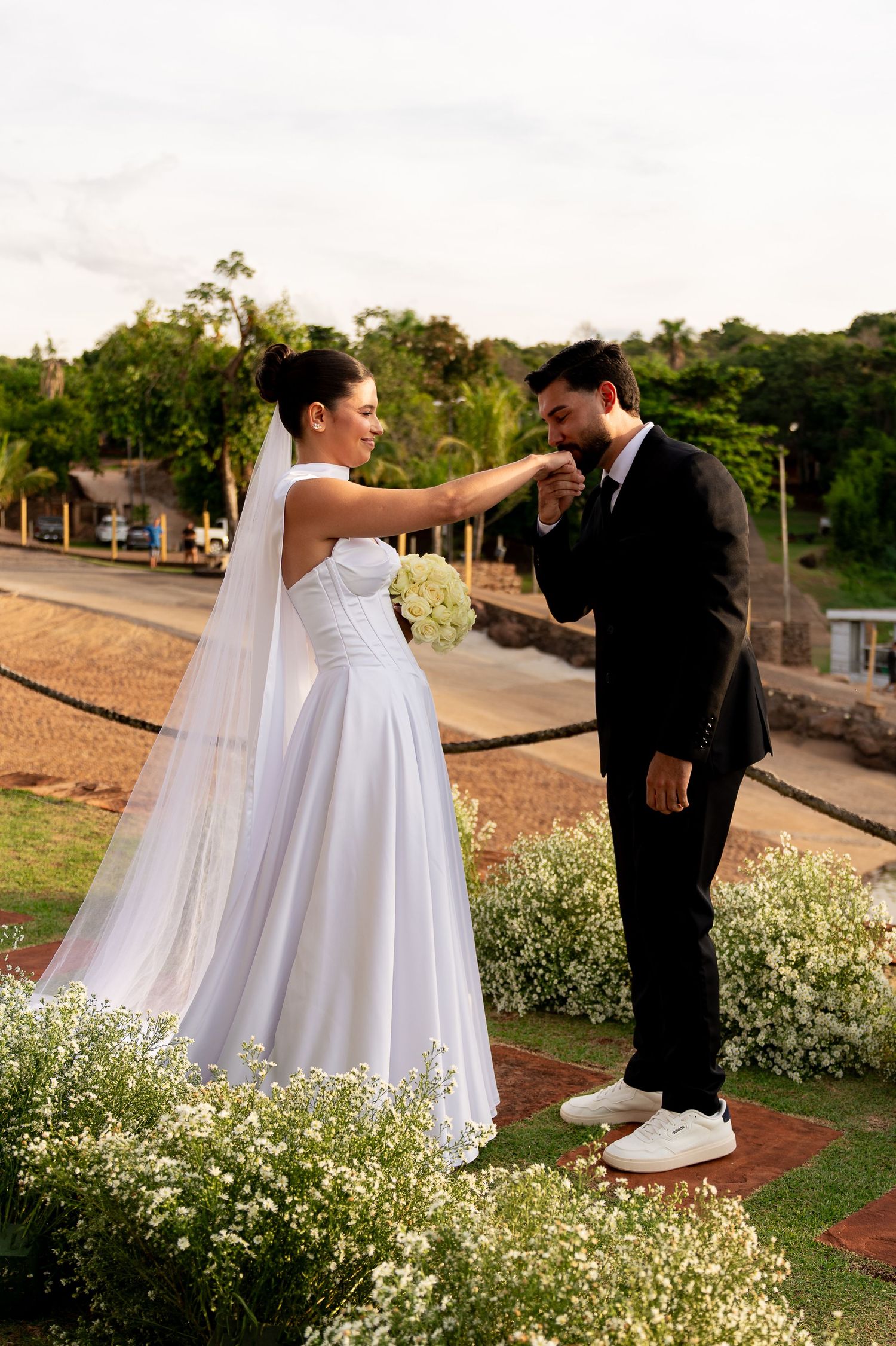 Groom in black suit kisses bride's hand outdoors, bride wearing white gown and veil holding bouquet.