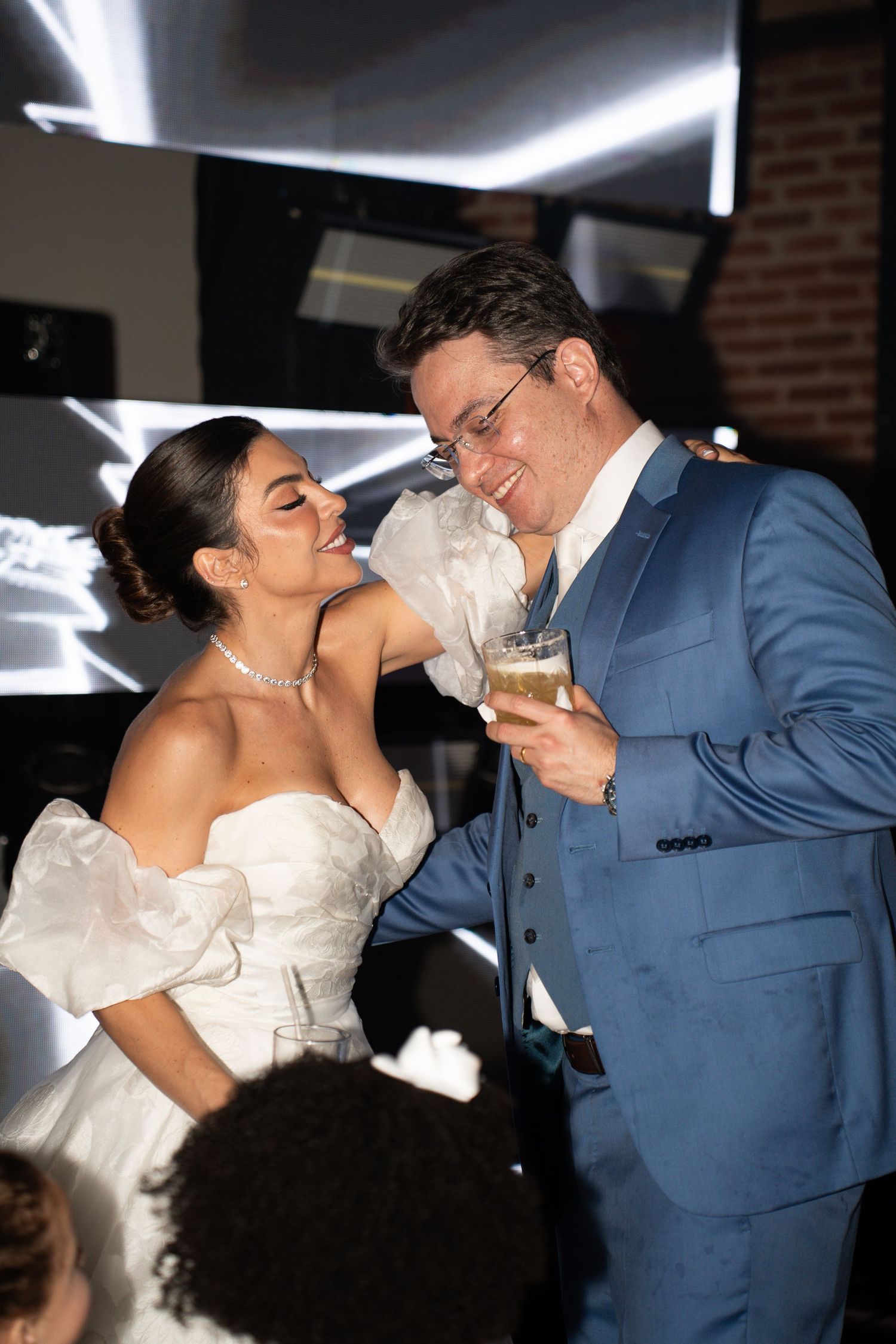 Bride in white off-shoulder gown and groom in blue suit share a joyful moment with drinks at their wedding reception.