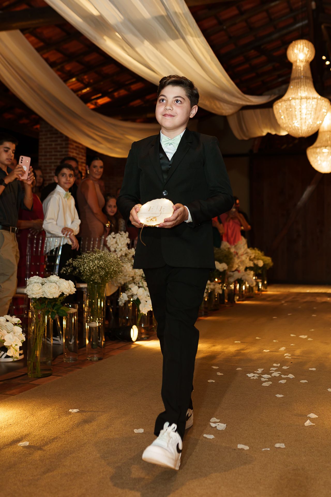 A young boy in a black suit carries a ring pillow down a decorated wedding aisle with floral arrangements and guests.