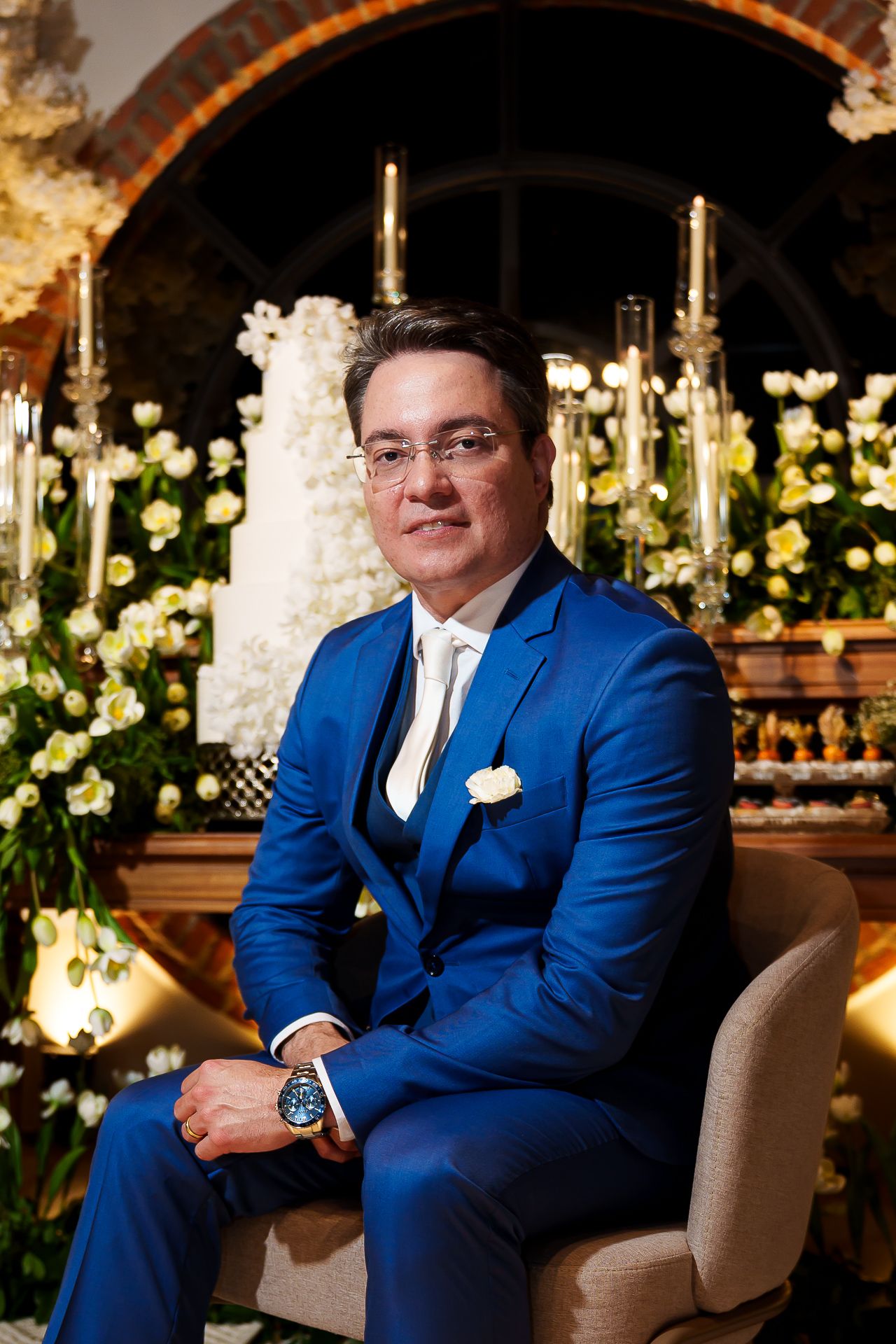 Man in blue suit seated at elegant wedding reception with white floral arrangements and candelabras.