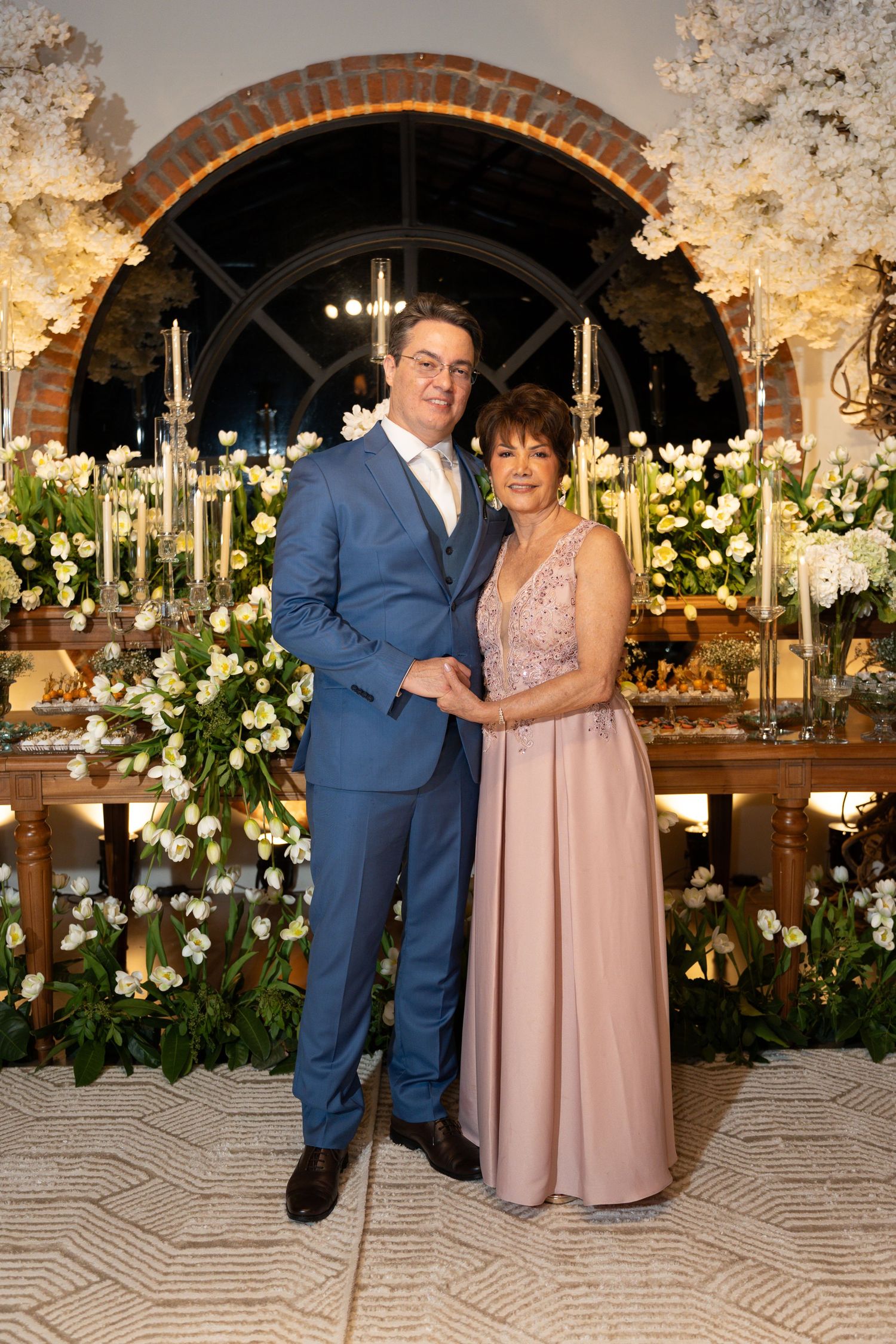 A man in a blue suit and woman in a pink gown pose together at an elegant floral wedding reception.