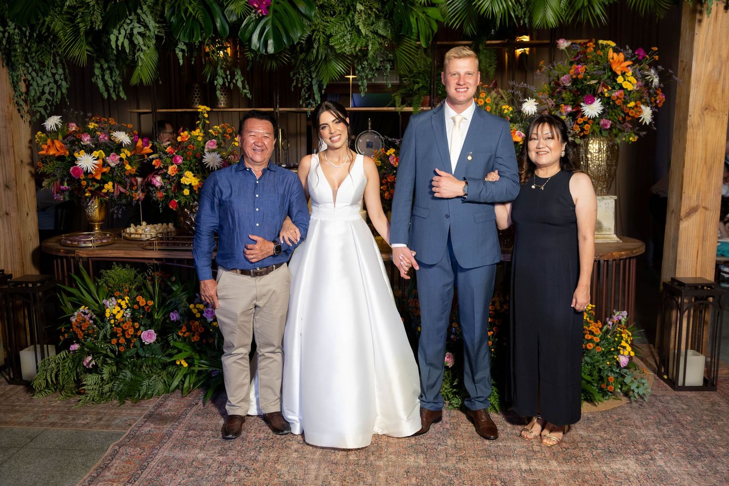 Bride in white gown poses with groom in blue suit and two guests amid lush floral backdrop at elegant wedding reception.
