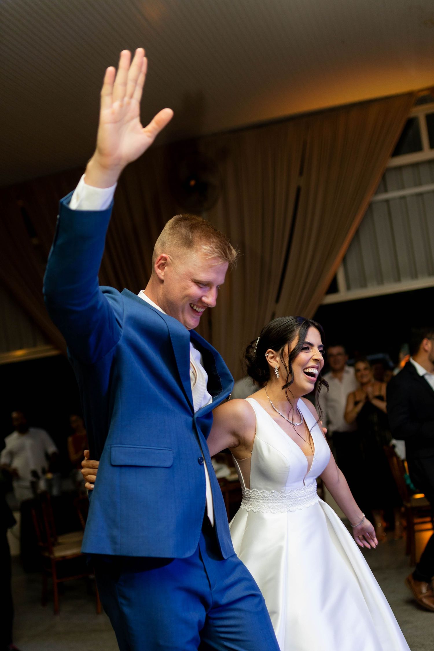Joyful bride and groom in blue suit entering wedding reception, groom waving to guests, bride laughing in white gown.