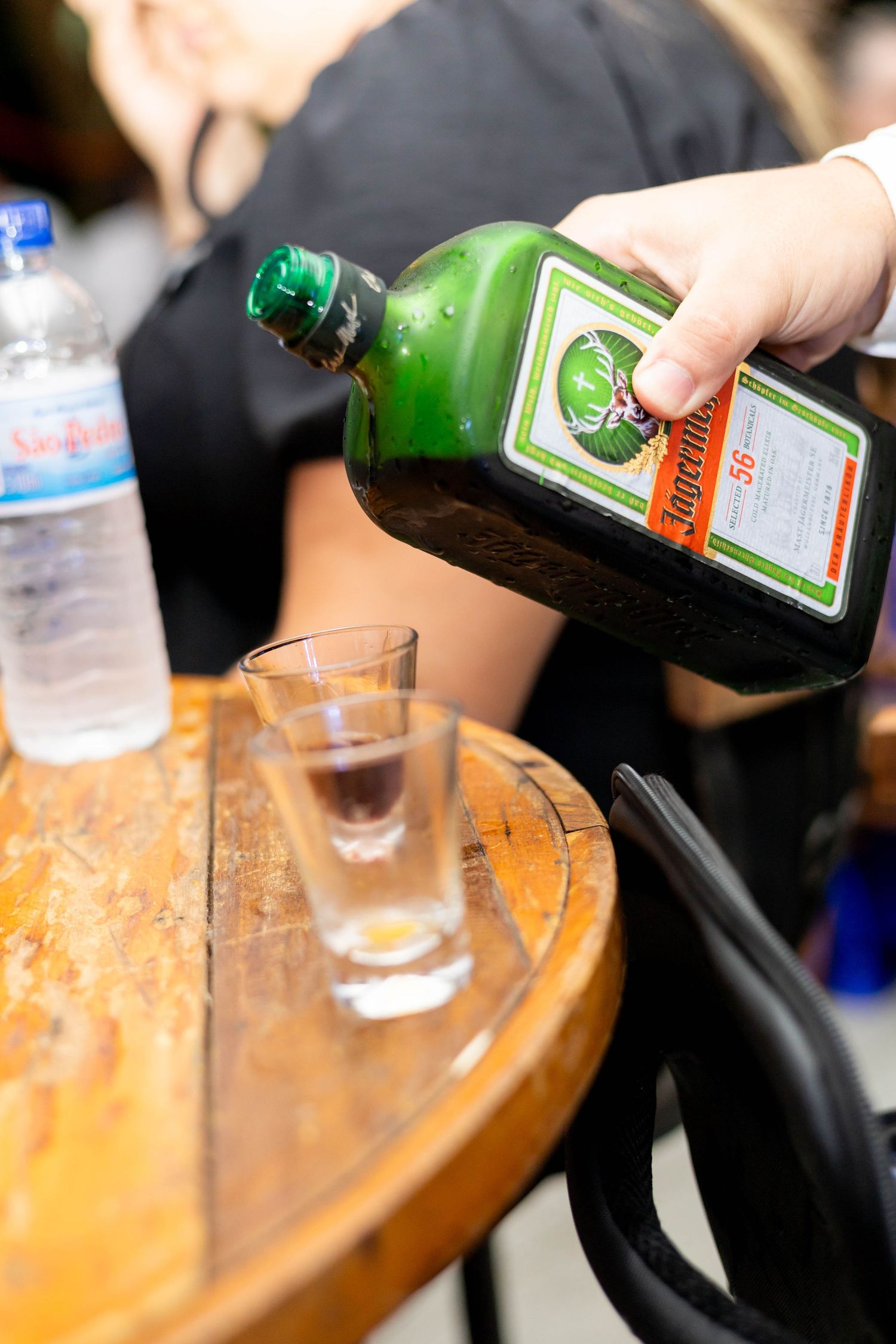 Hand pouring Jagermeister from a green bottle into a shot glass on a wooden table.
