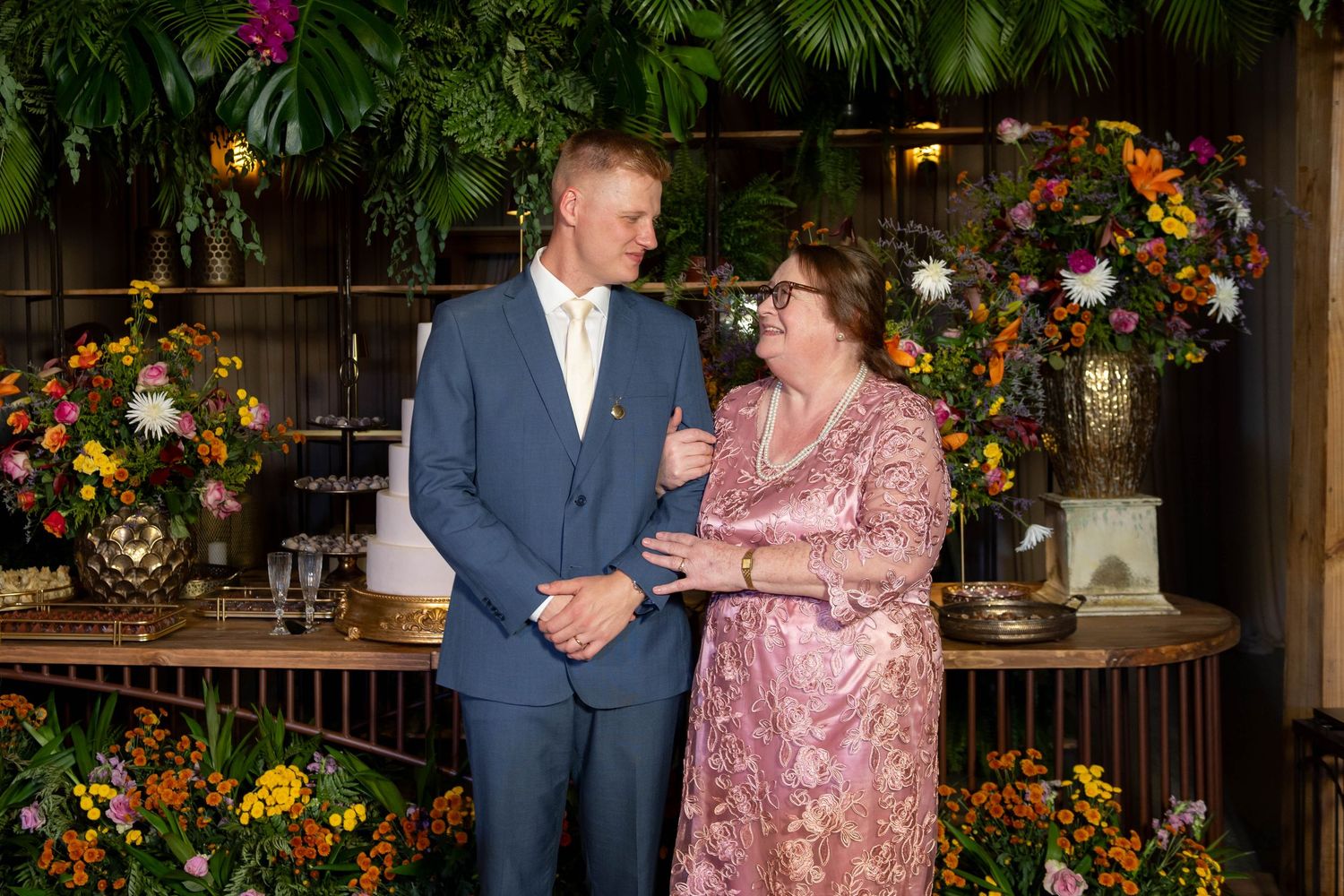 Couple in formal attire smiling together at an elegant floral event venue with colorful arrangements.