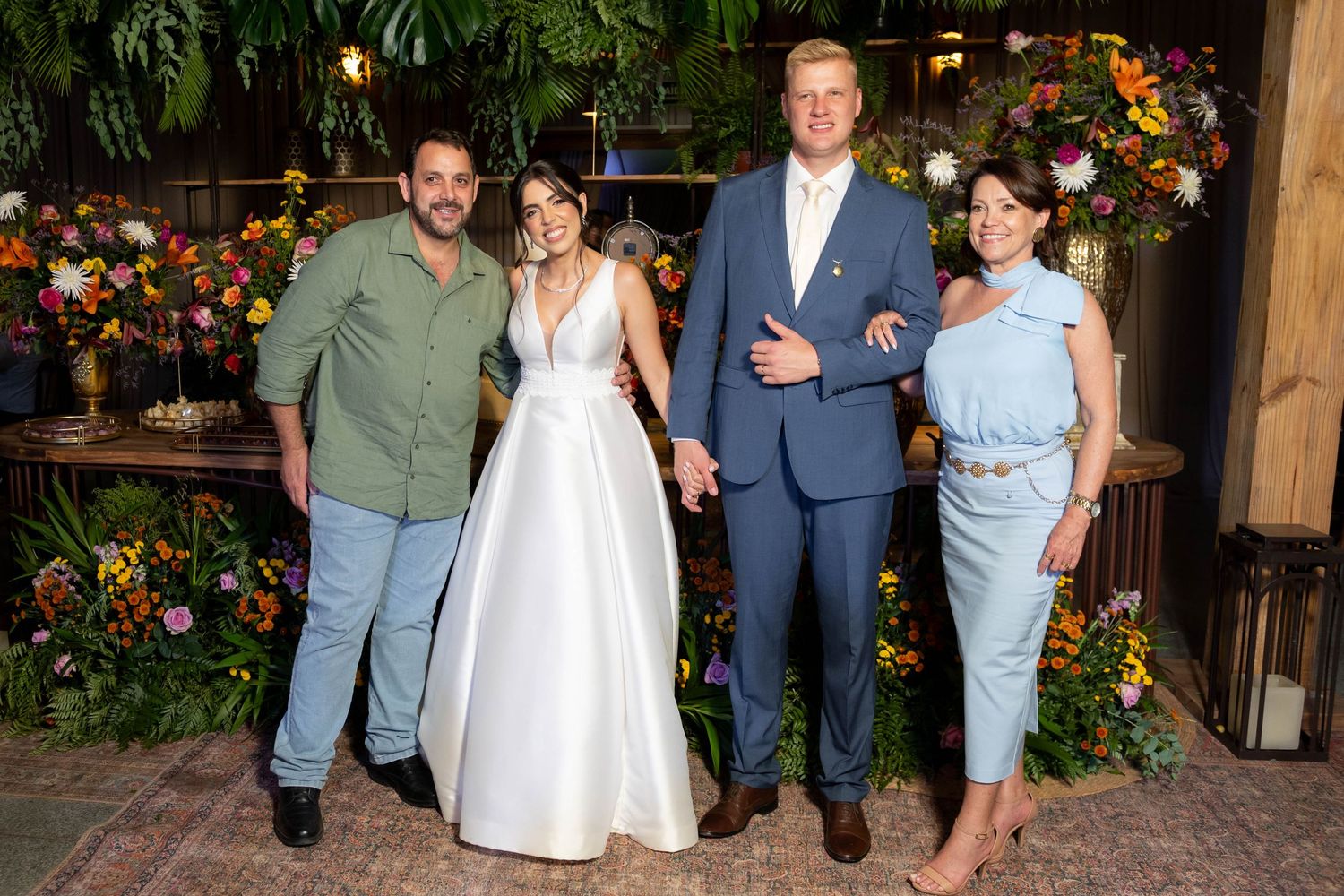 Two couples pose together at a floral wedding reception, one in a white bridal gown and the other in elegant formal attire.