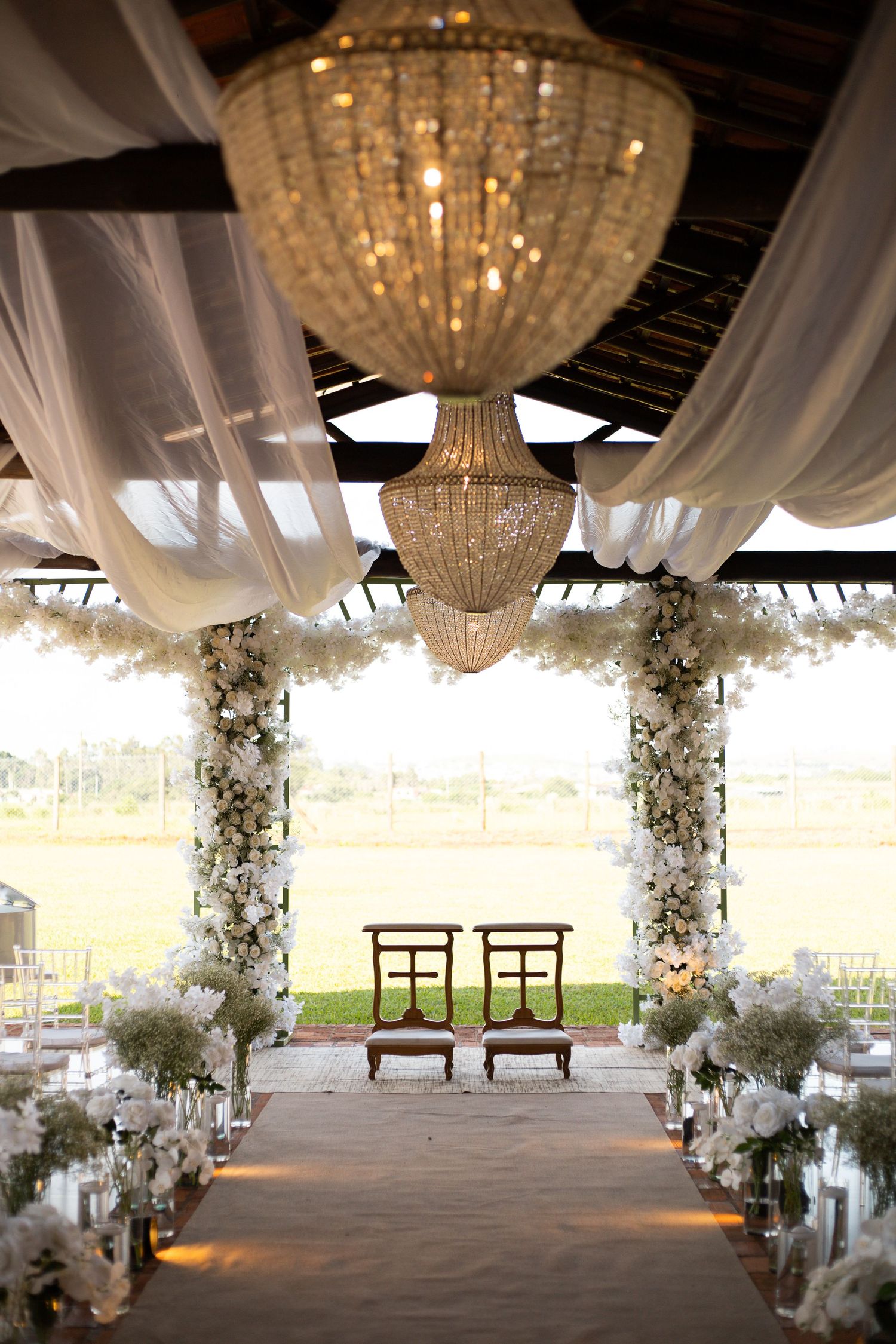 Elegant wedding ceremony aisle with crystal chandeliers, white floral arches, draped fabric, and two wooden chairs at the altar.