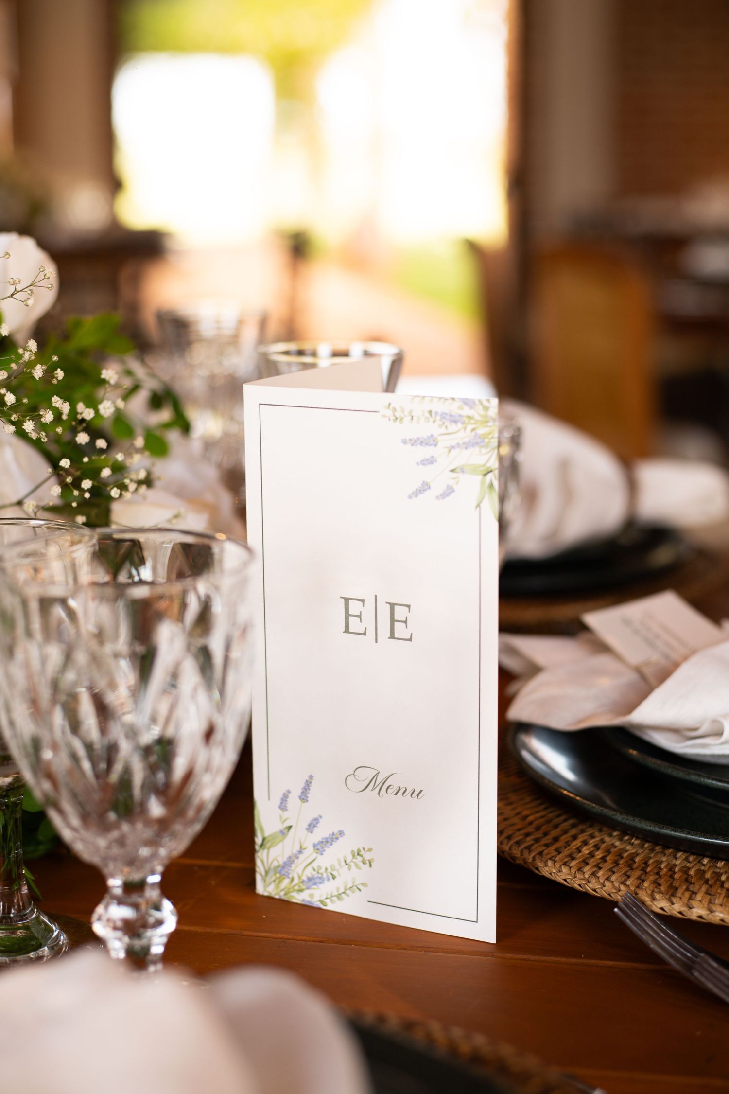 Elegant wedding menu card with monogram E|E, floral accents, crystal glassware, and black plates on a wooden table.