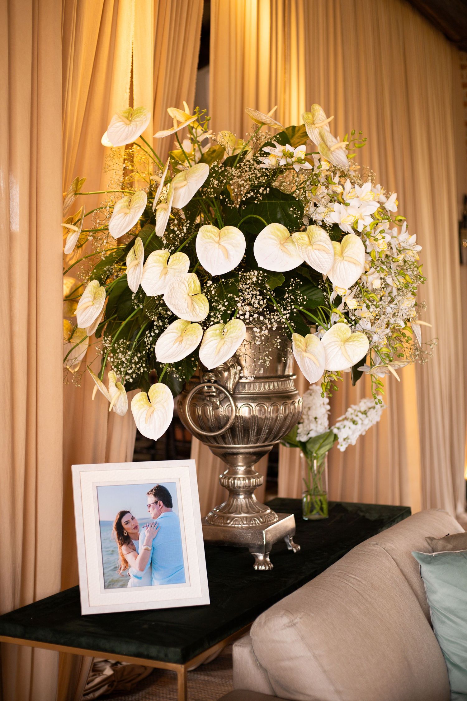 Elegant silver urn vase with white anthuriums and greenery beside a framed couple photo on a dark table.