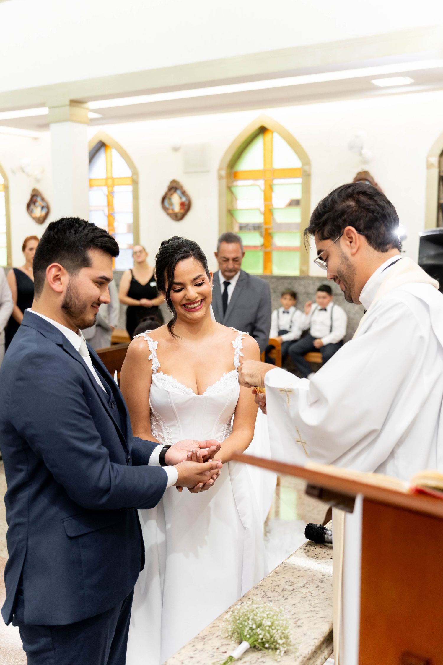 A smiling bride and groom exchange rings at a church altar, officiated by a priest during their wedding ceremony.