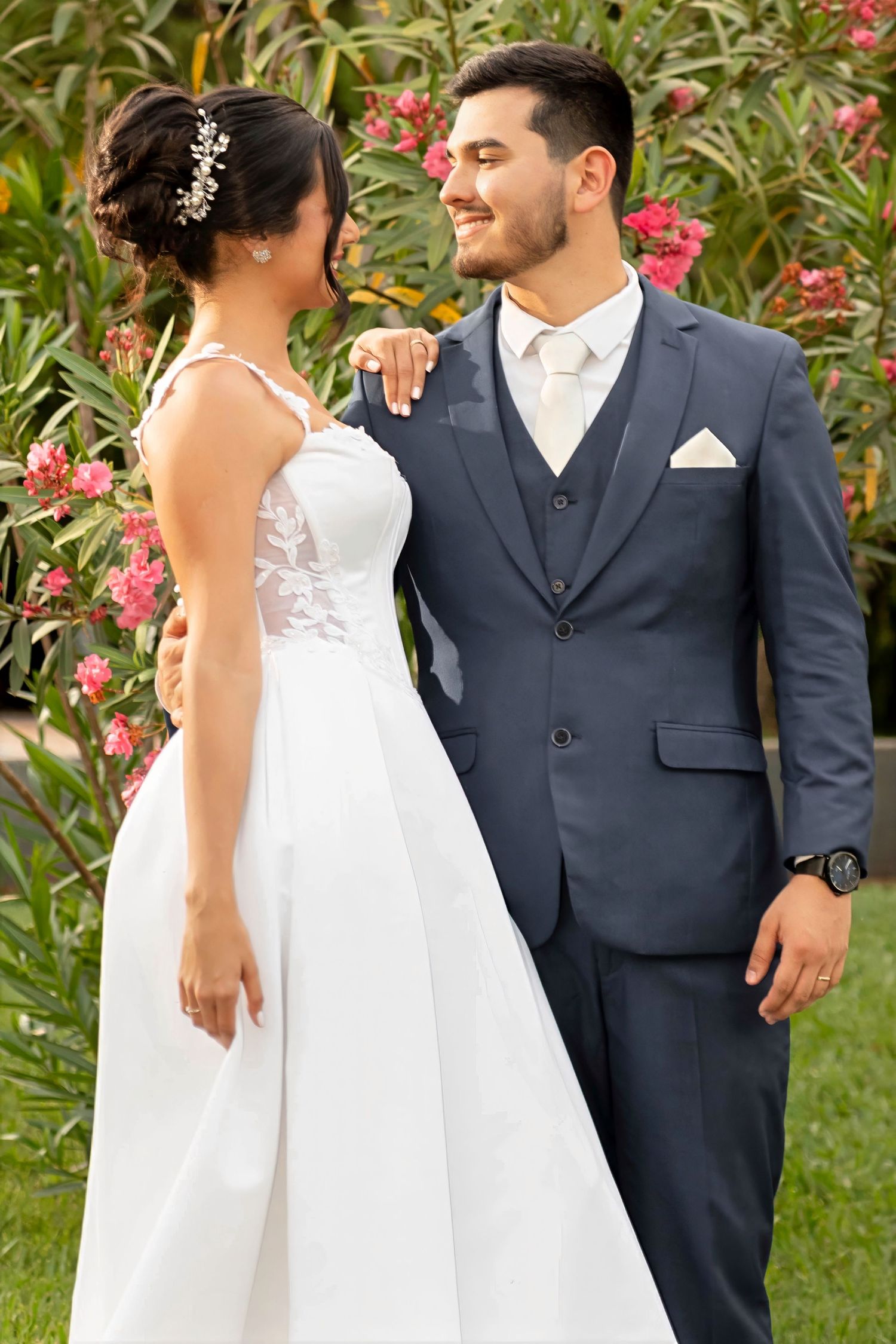 Bride in white lace gown and groom in navy suit smiling at each other outdoors with pink flowers.