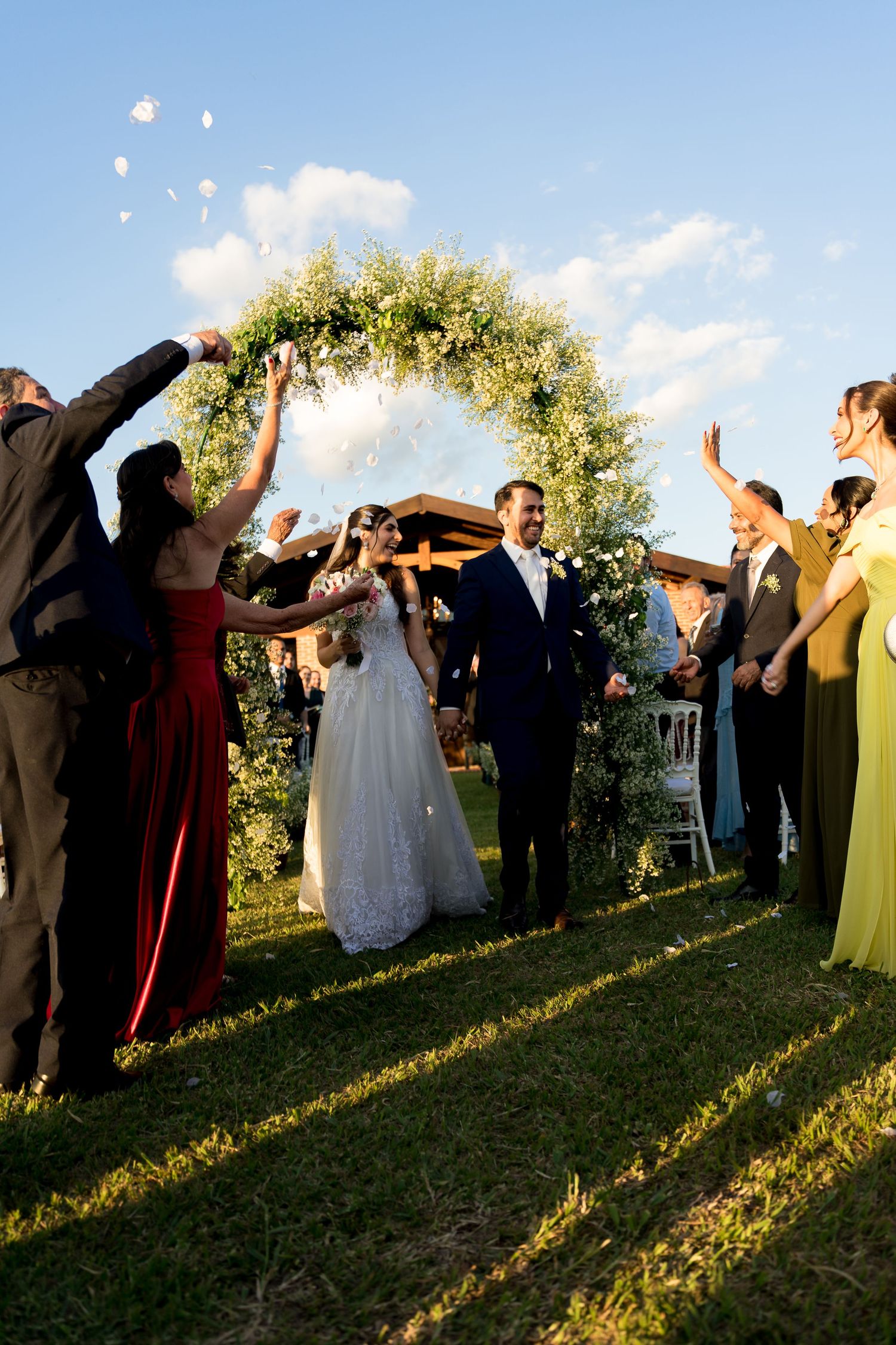Bride and groom walk through floral arch as guests toss petals at outdoor wedding ceremony on sunny day.