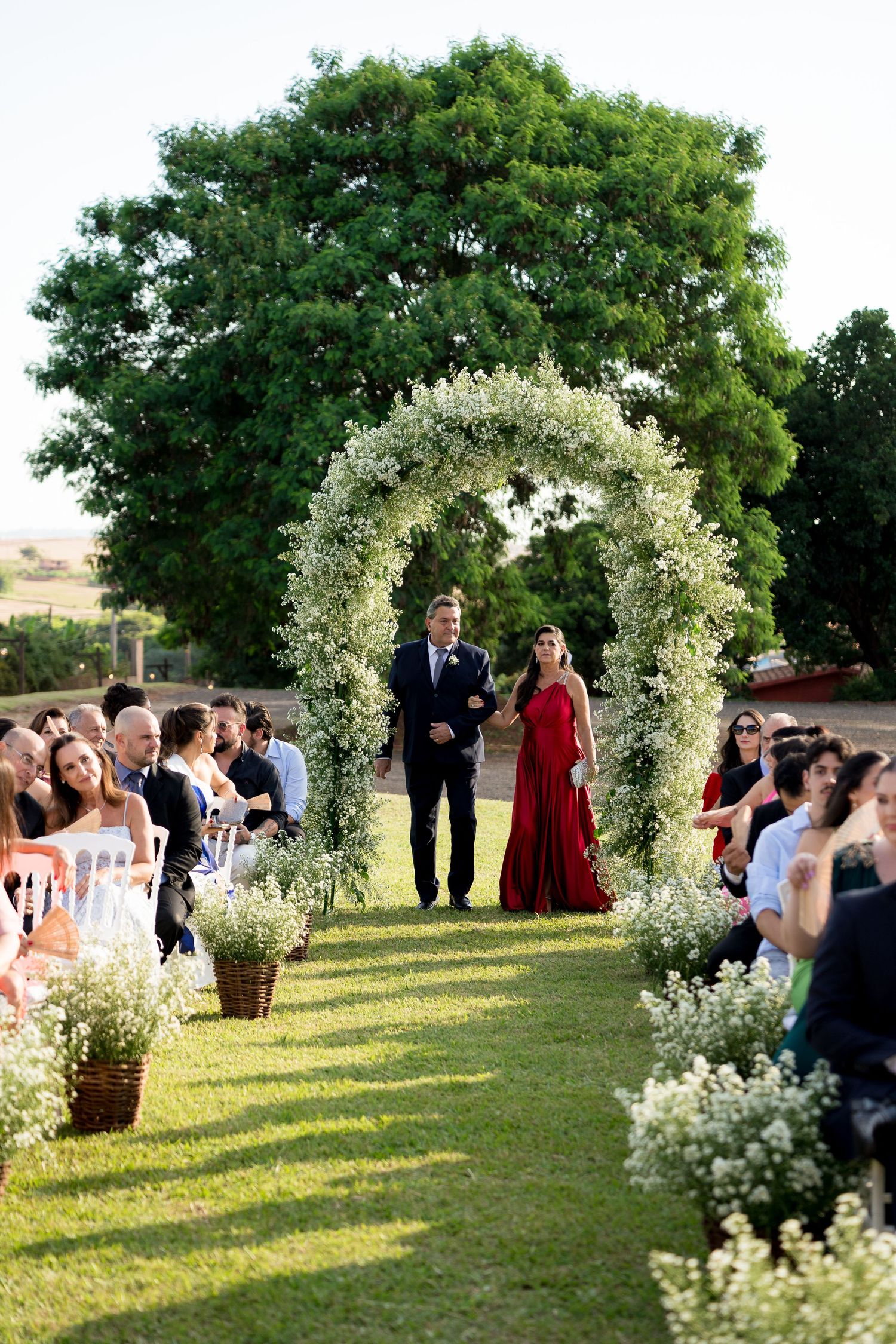 Outdoor wedding ceremony with floral arch, couple walking down aisle lined with white flowers and seated guests.