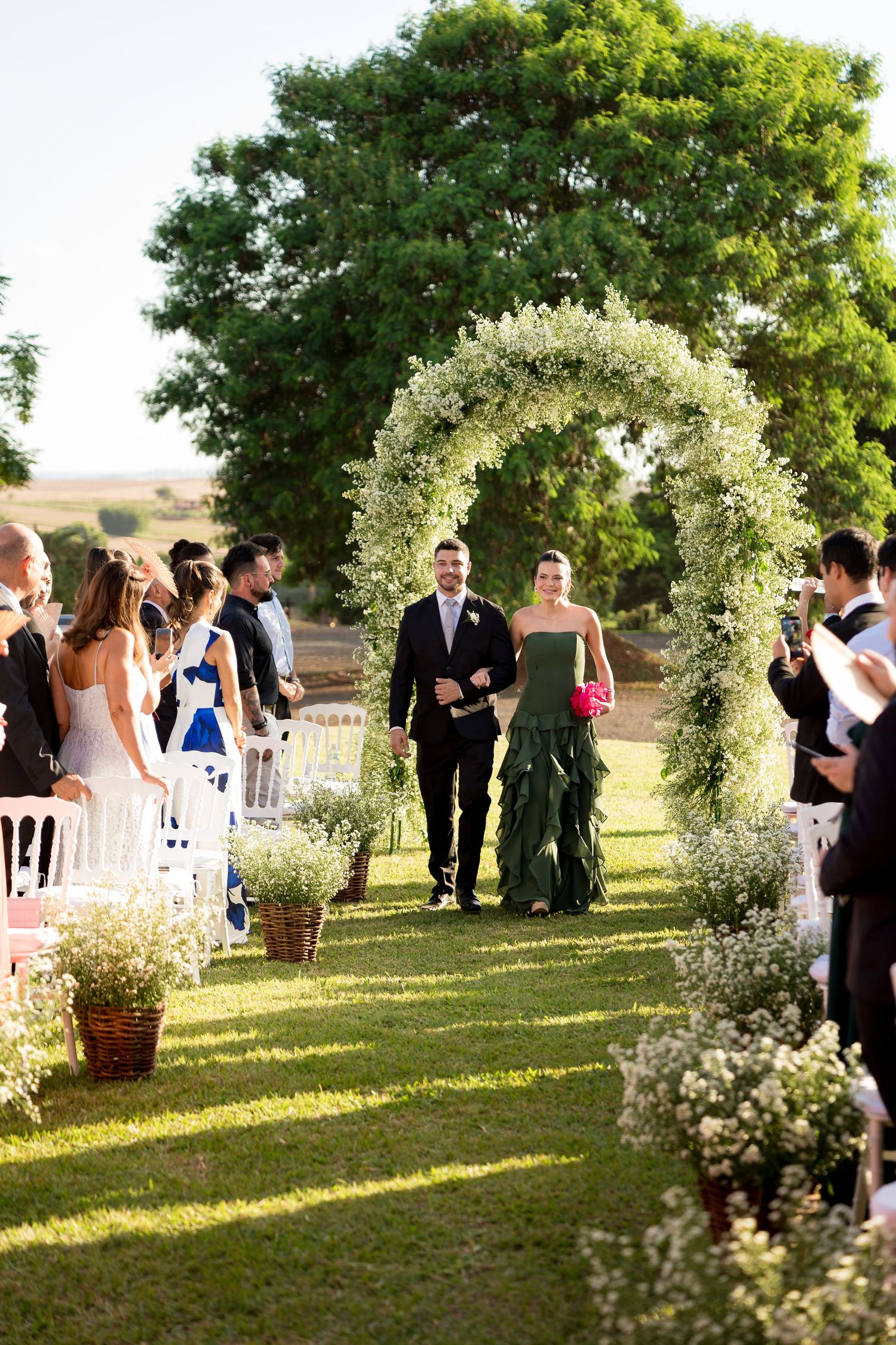 Couple walks down outdoor wedding aisle under floral arch, guests seated on white chairs, lush greenery backdrop.
