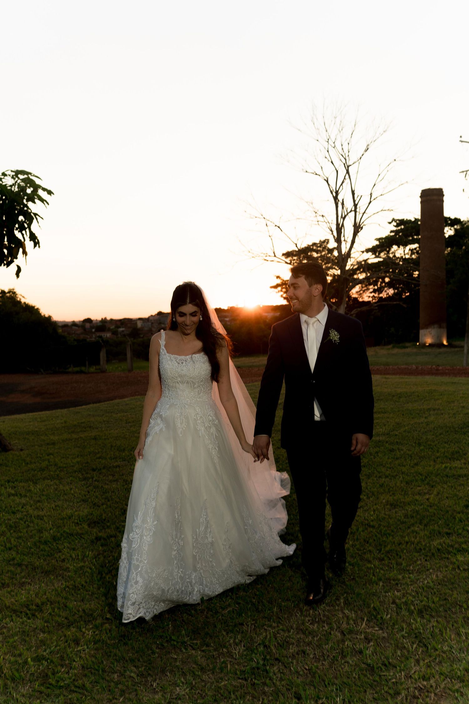 Bride and groom walk hand-in-hand on green lawn at golden sunset, she in lace ballgown, he in black tuxedo.