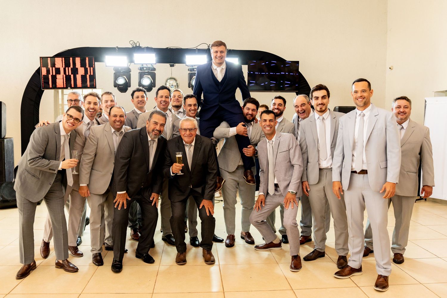 Large group of men in gray suits posing together at a wedding, with the groom being lifted up in celebration.