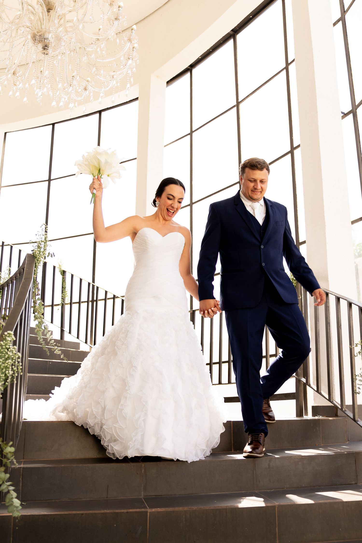 Bride in white gown and groom in navy suit walk down stairs holding hands, bride raising bouquet joyfully.