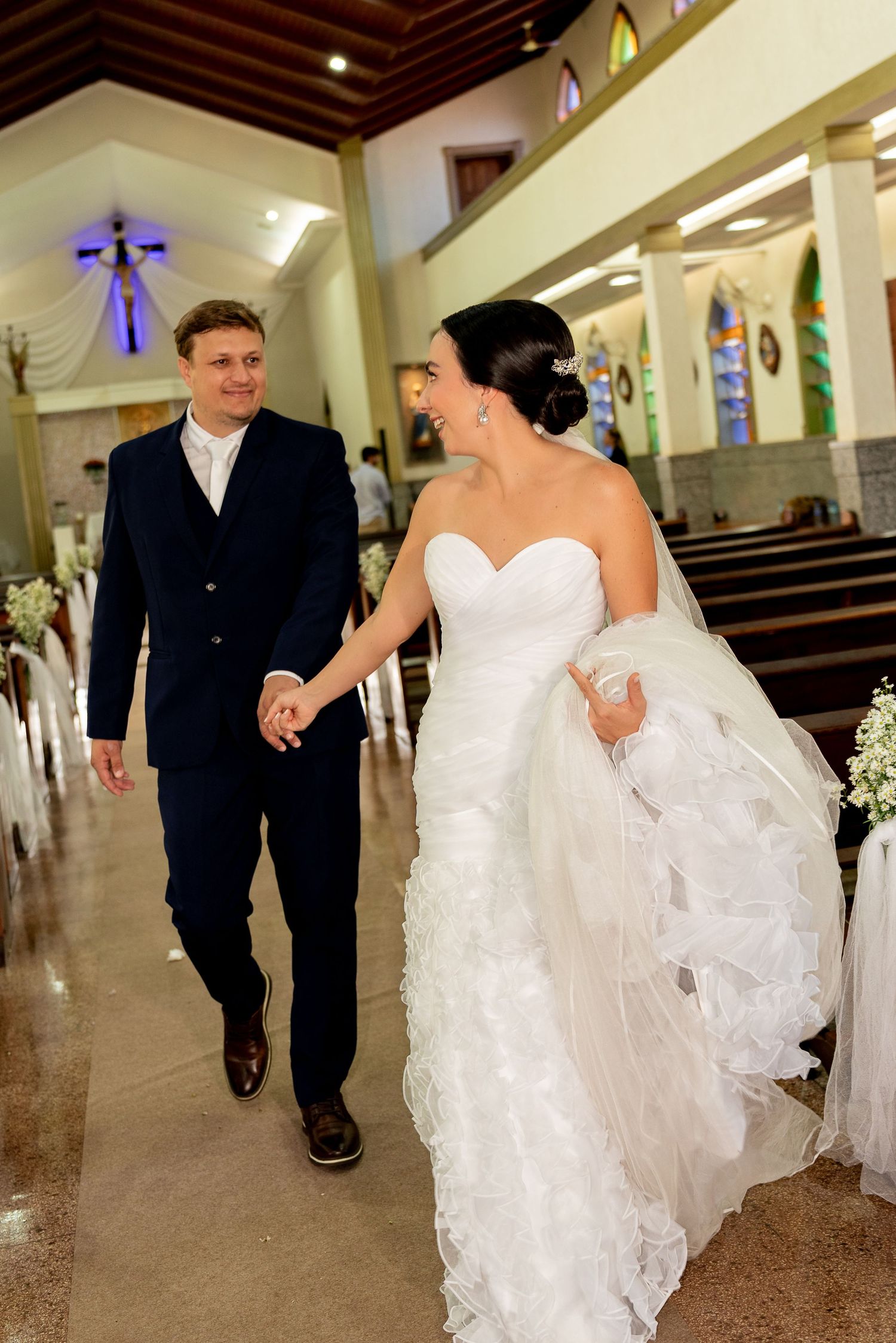 Newlywed couple walking hand in hand down church aisle, bride in white ruffled gown and groom in navy suit.