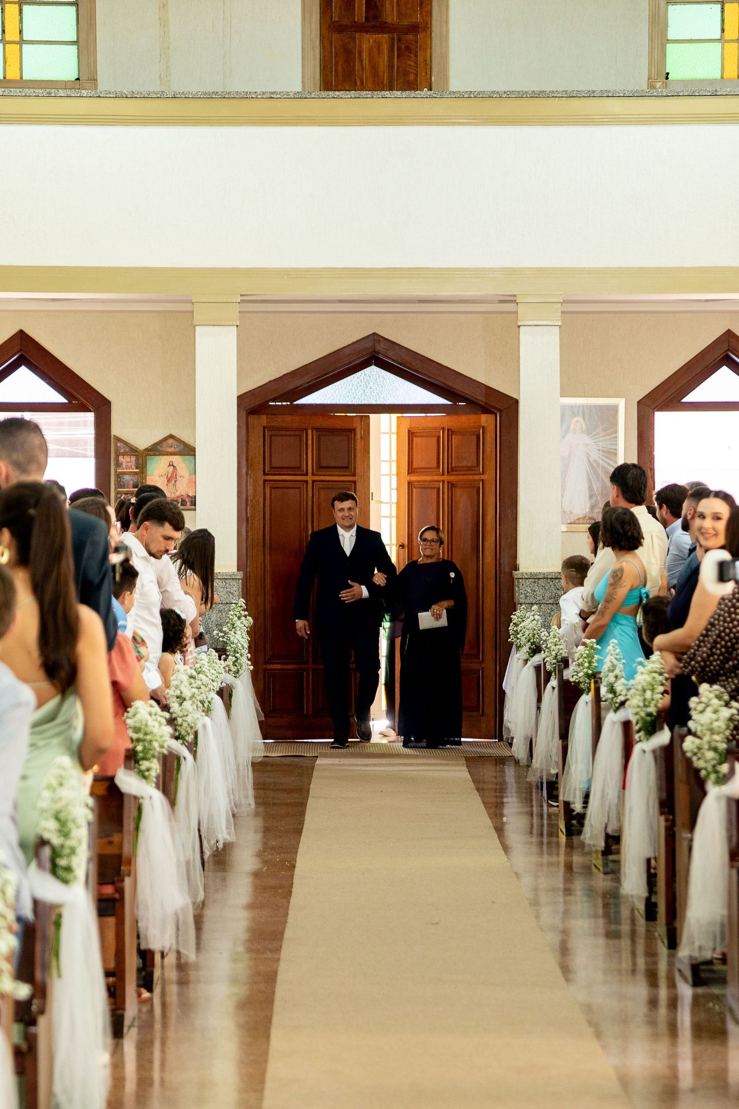 A groom walks down the church aisle with a guest during a wedding ceremony, with pews decorated in white tulle.
