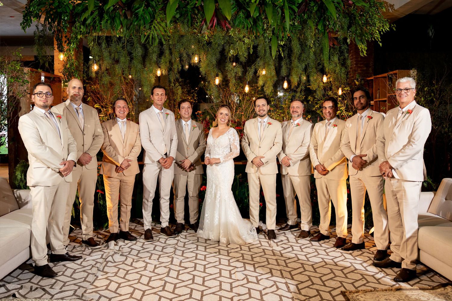 Wedding party poses indoors, bride in white gown flanked by groomsmen in tan suits under lush greenery with warm Edison lights.