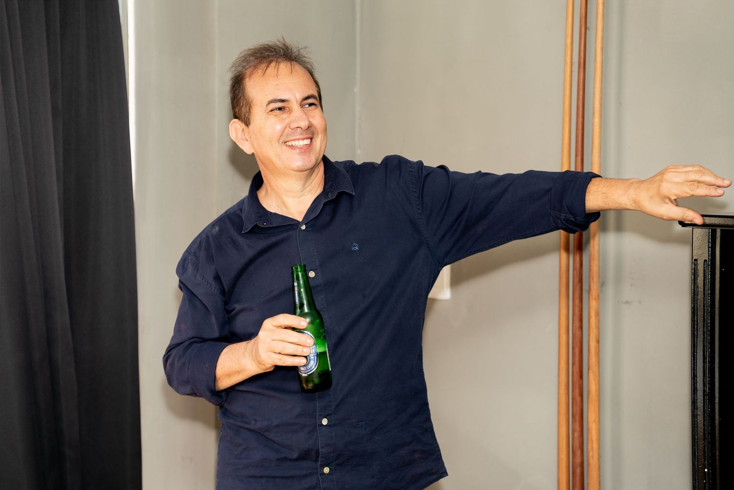 Smiling man in navy shirt holding a green beer bottle, leaning against a wall at a casual indoor event.