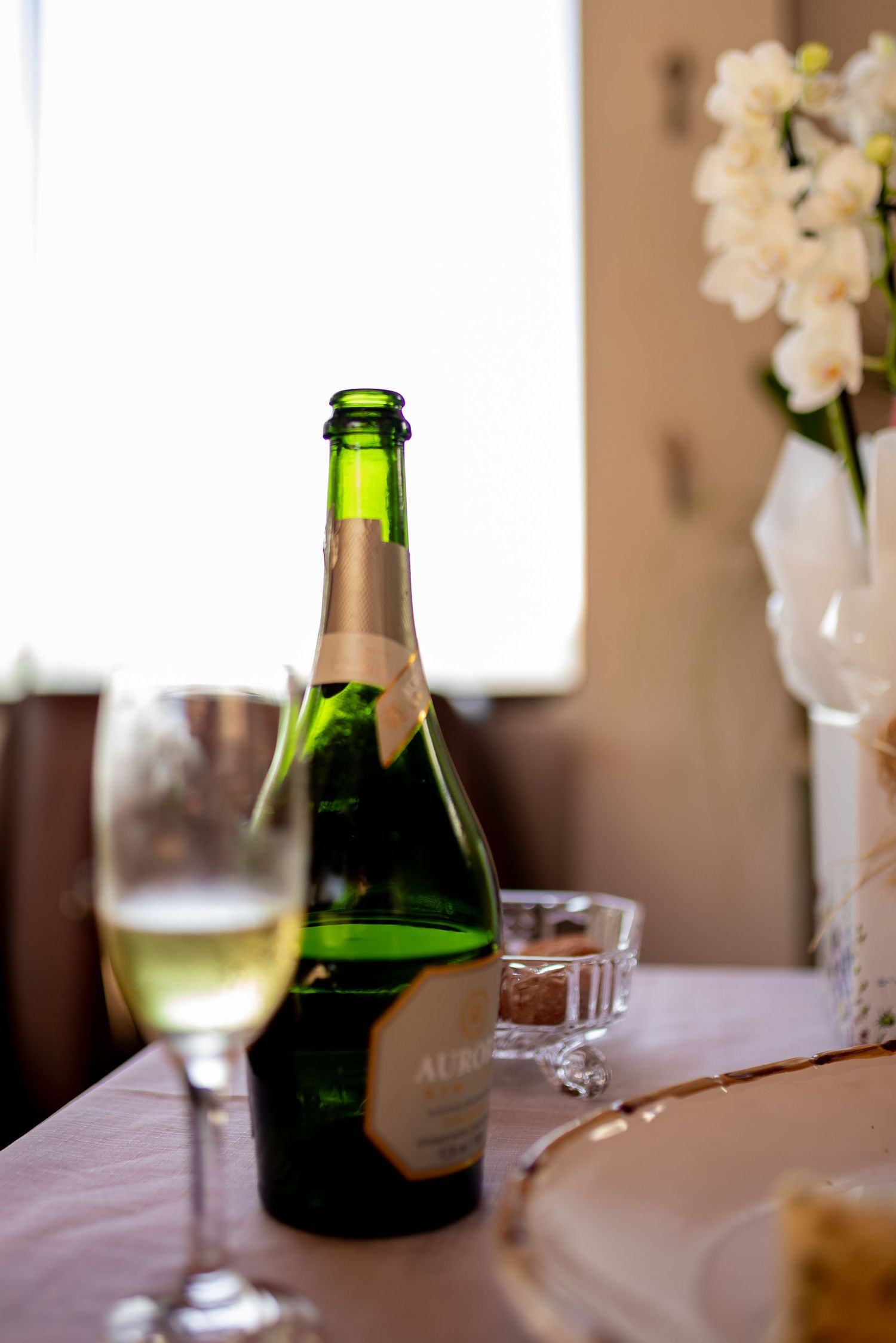 Green champagne bottle with a glass of white wine on a table, with white flowers in a vase in the background.