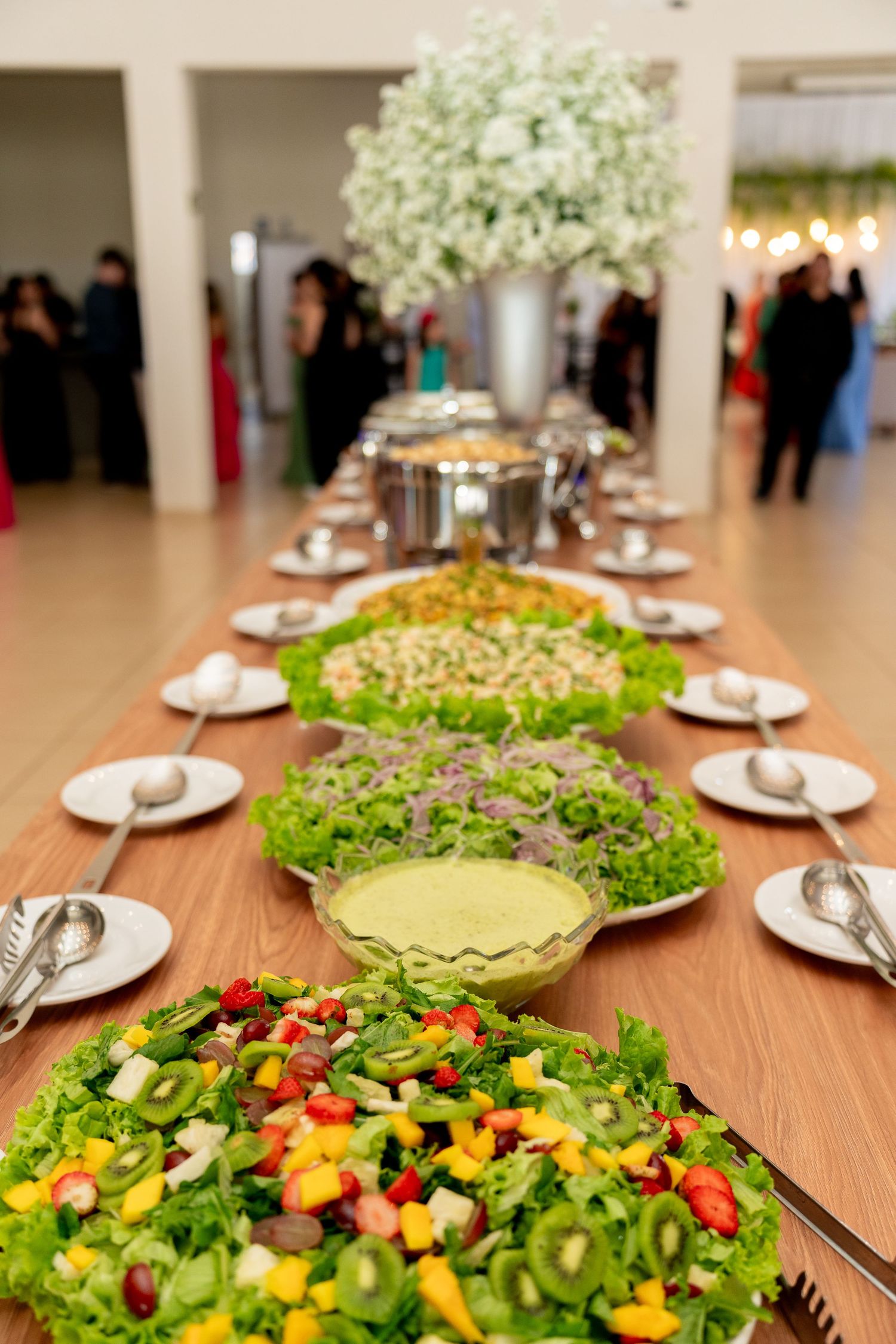 Elegant buffet table with colorful fresh salads, fruit, and greens at a formal event with floral centerpiece.