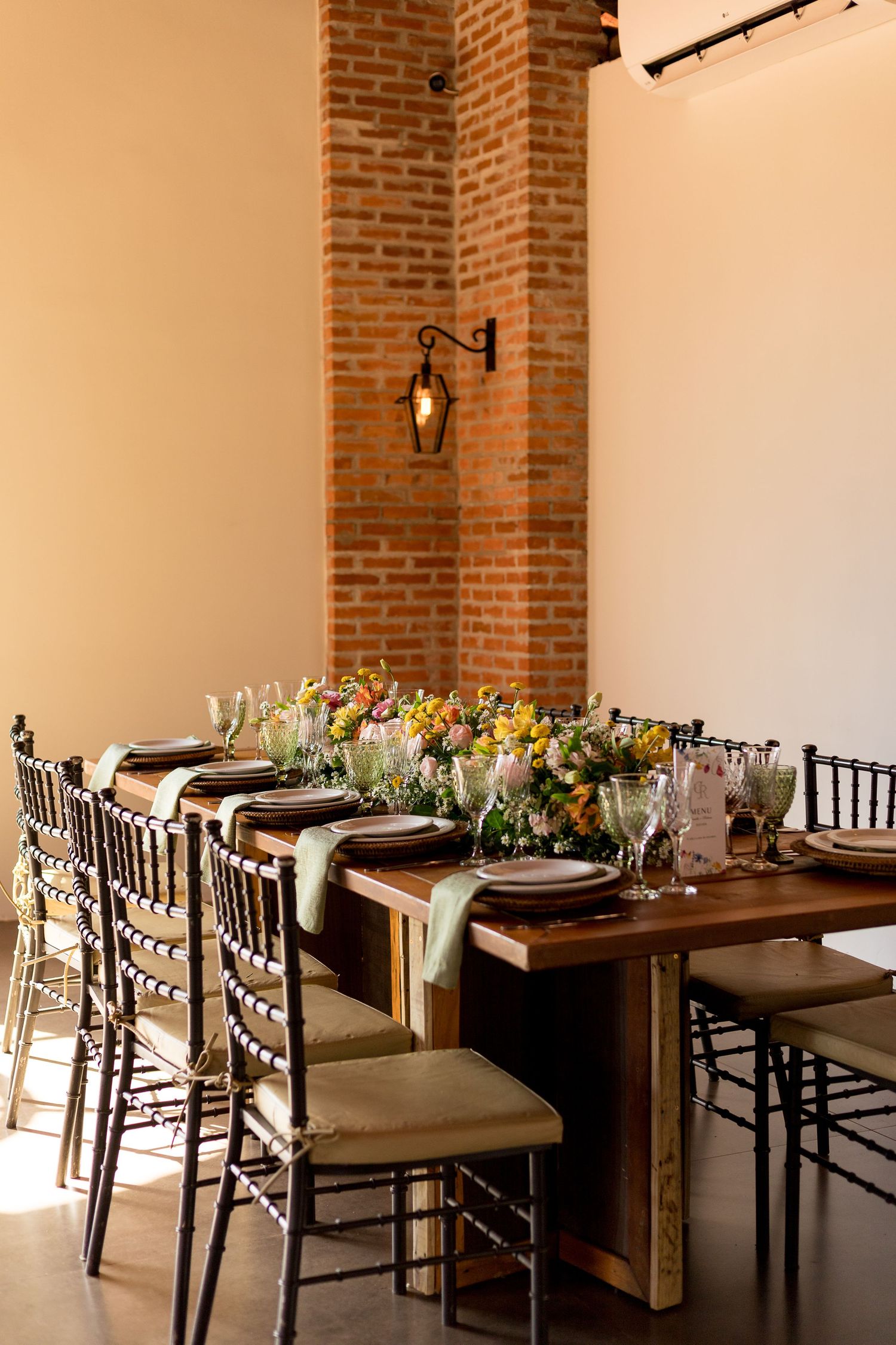 Elegant rustic dining table with floral centerpiece, chiavari chairs, and exposed brick column in a warmly lit event space.