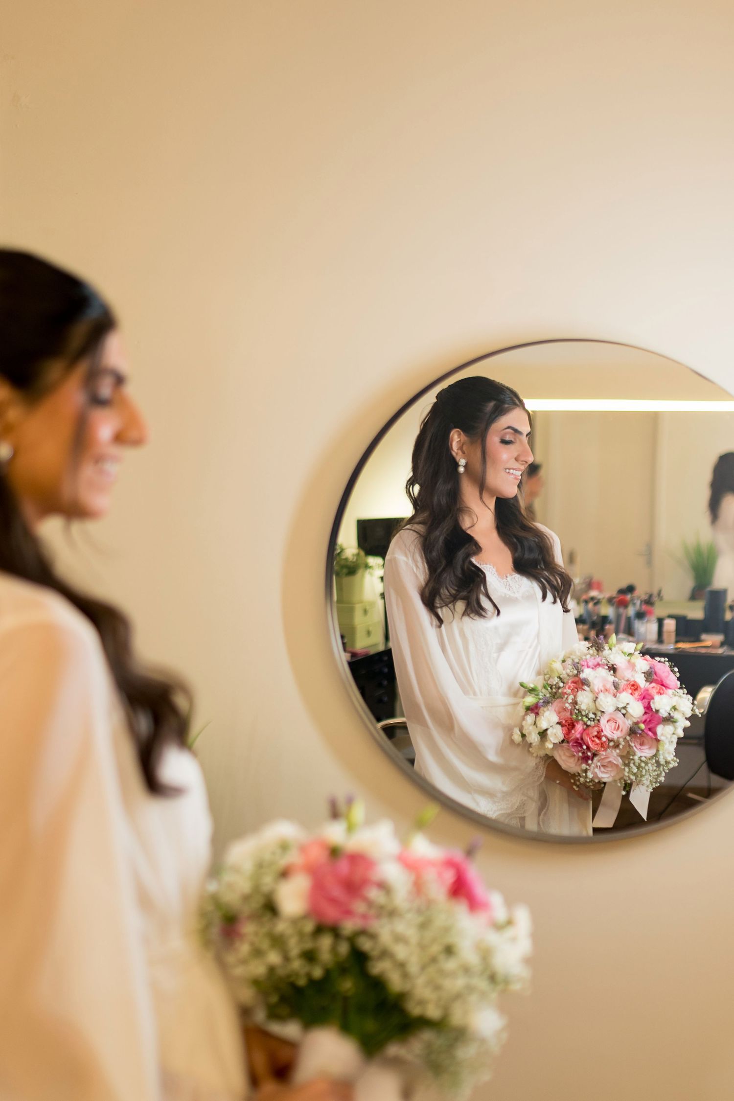 Bride in white gown holds pink bouquet, smiling reflection visible in round mirror during wedding preparations.