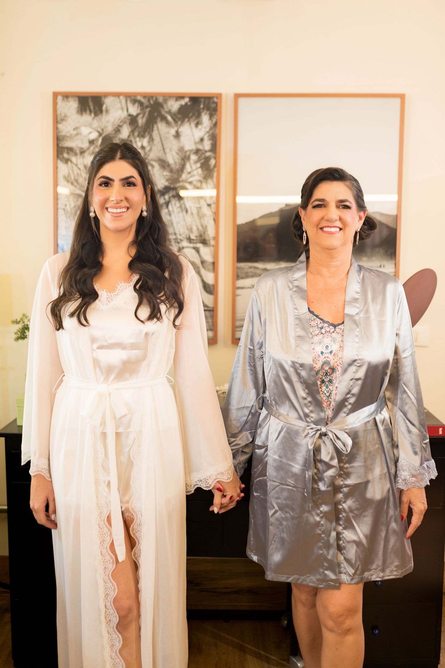 Two women in satin robes holding hands and smiling, one in white bridal robe, one in silver, standing before framed artwork.