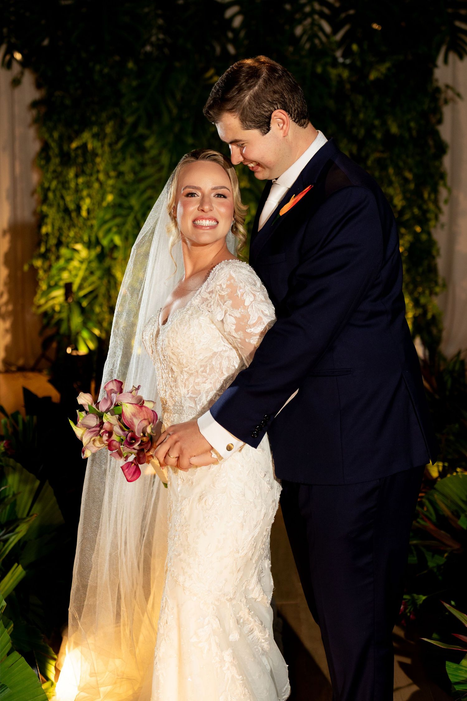 Bride in lace gown holding floral bouquet smiles as groom in navy suit with orange tie embraces her at evening wedding ceremony.